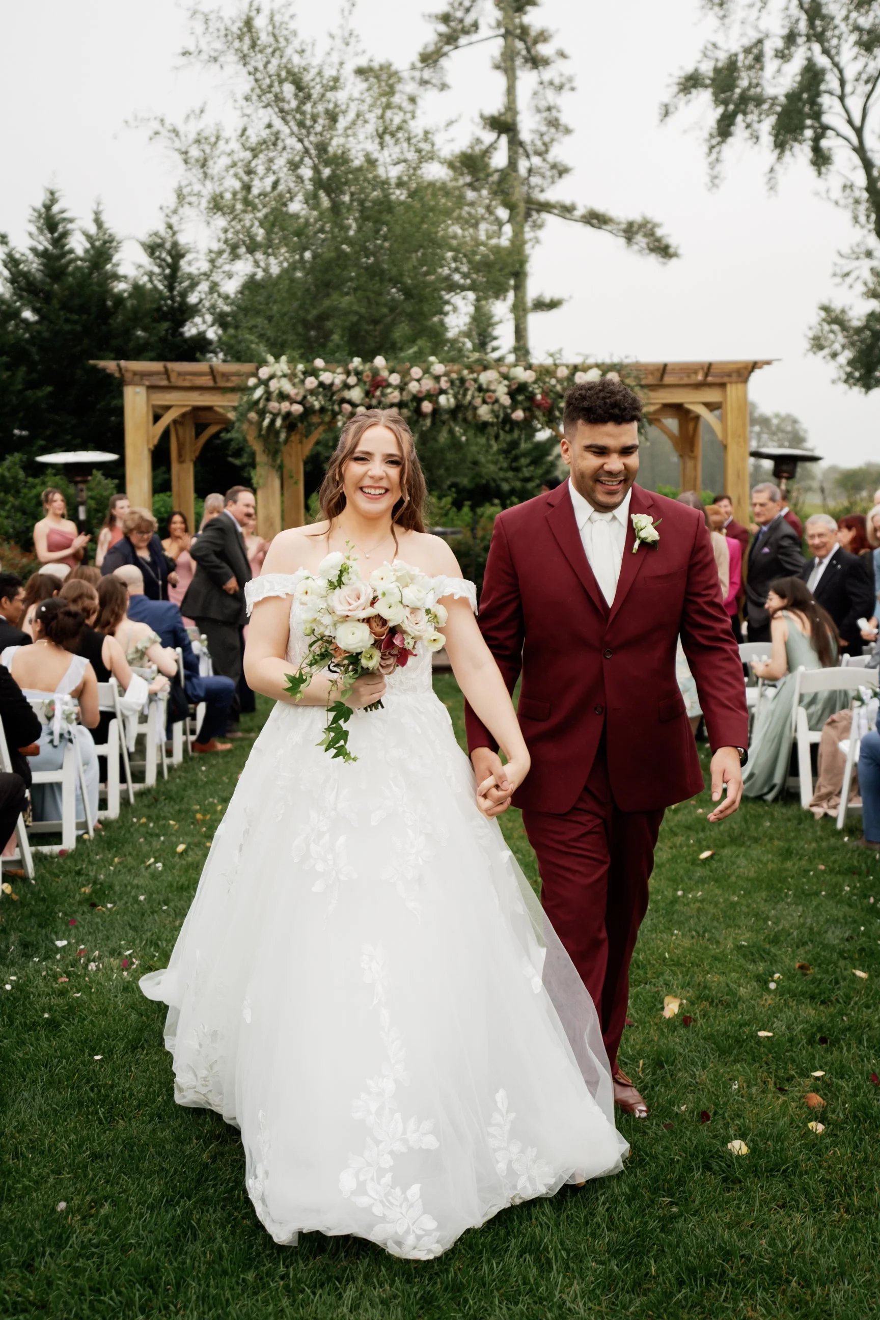 A newlywed couple walking hand in hand outdoors at their wedding ceremony. The bride is wearing a white off-the-shoulder lace wedding gown and holding a bouquet, while the groom is in a maroon suit with a white shirt and boutonniere. Guests are seated and standing behind them, and there is a decorated wooden arch in the background surrounded by trees.