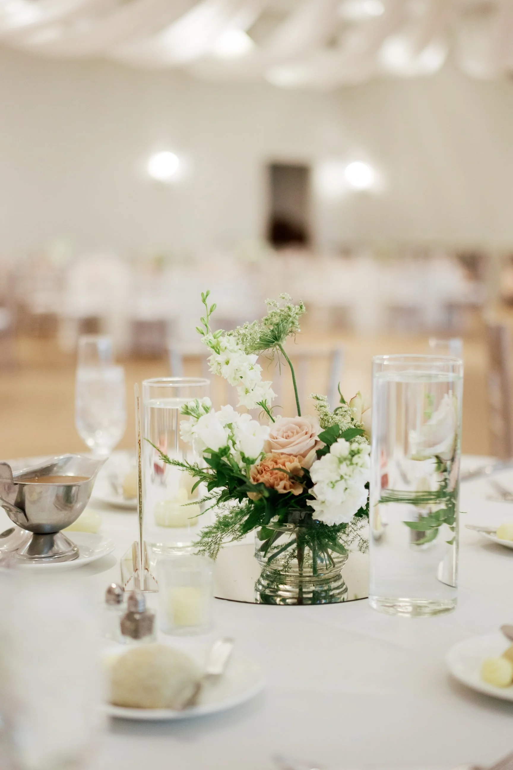 A close-up photo of a table centerpiece with white and blush pink flowers in a glass vase, with various drinking glasses and tableware around it, set in a bright, elegant event space.