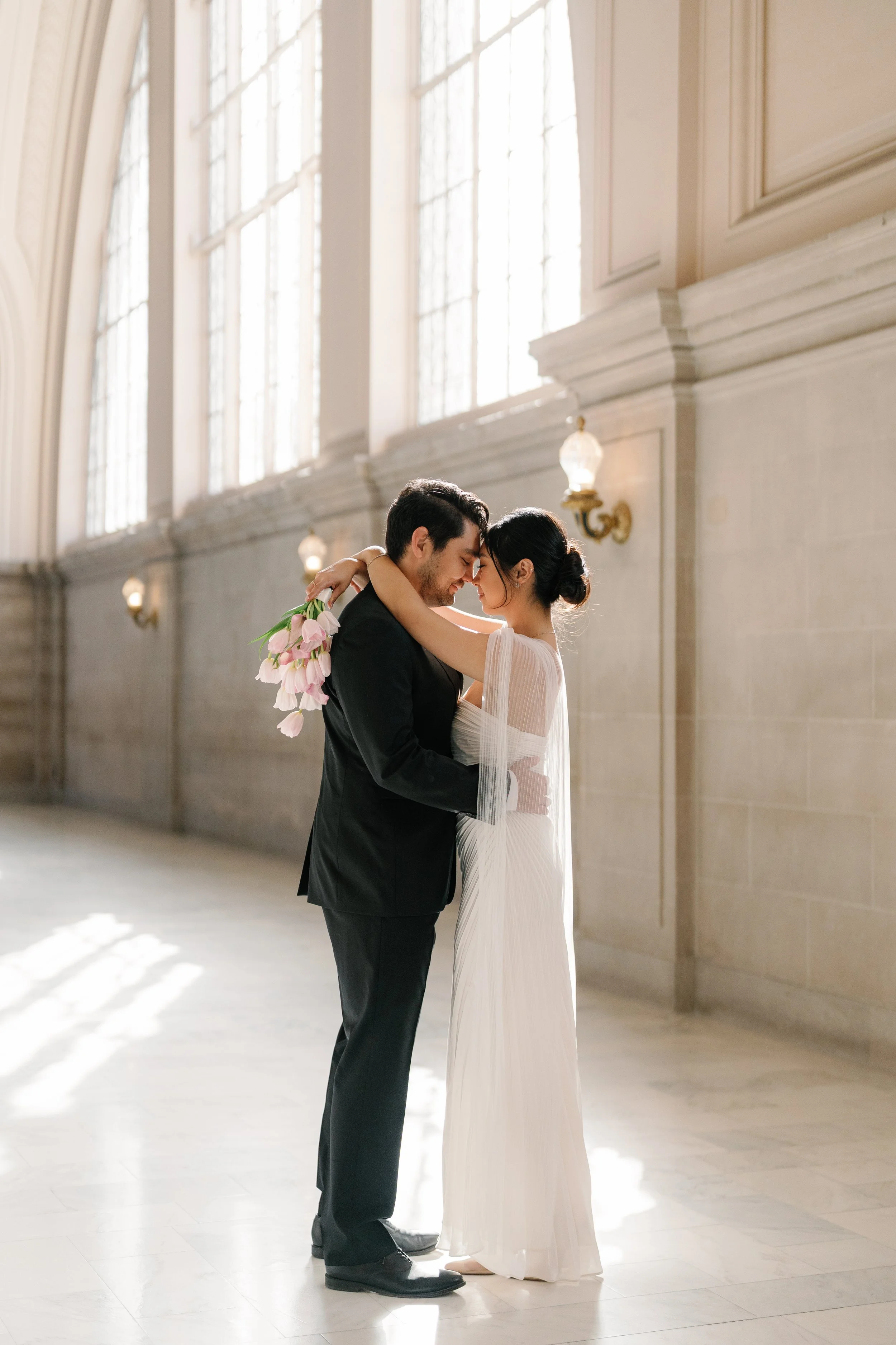 A couple dressed in wedding attire embracing each other in a grand hall with tall windows and soft lighting.