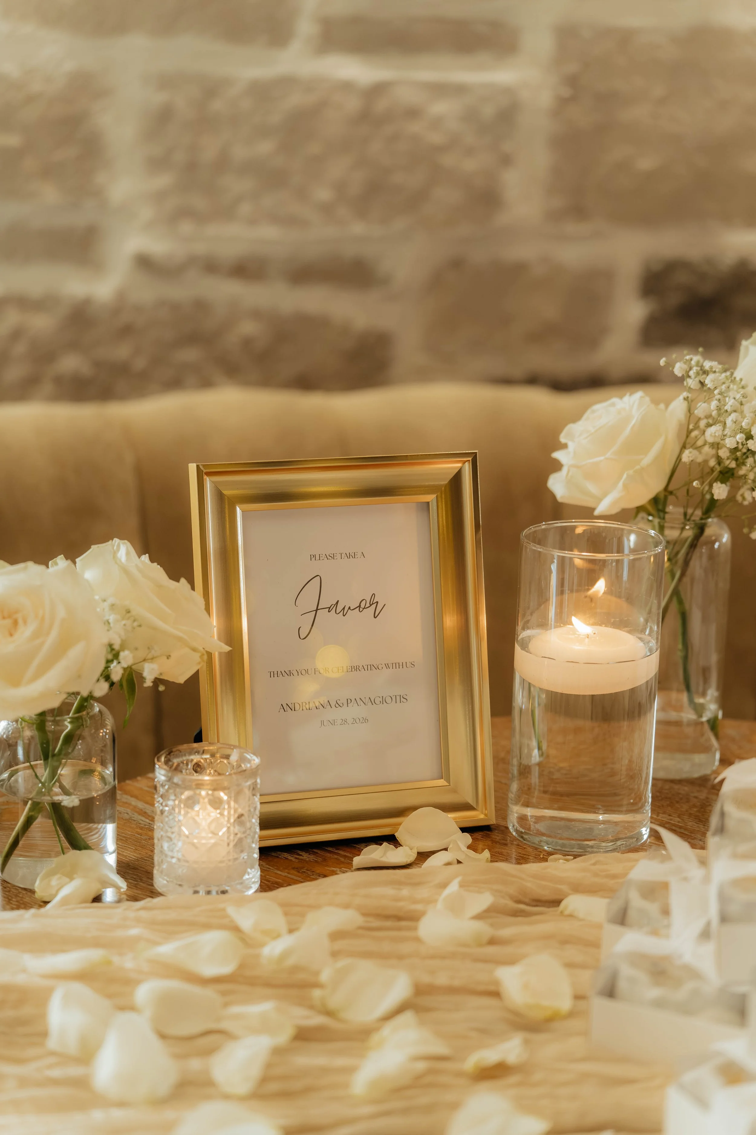 A wedding reception table decorated with white roses, candles, and a gold-framed sign with the names Andreina and Panagiotis, date June 28, 2026.