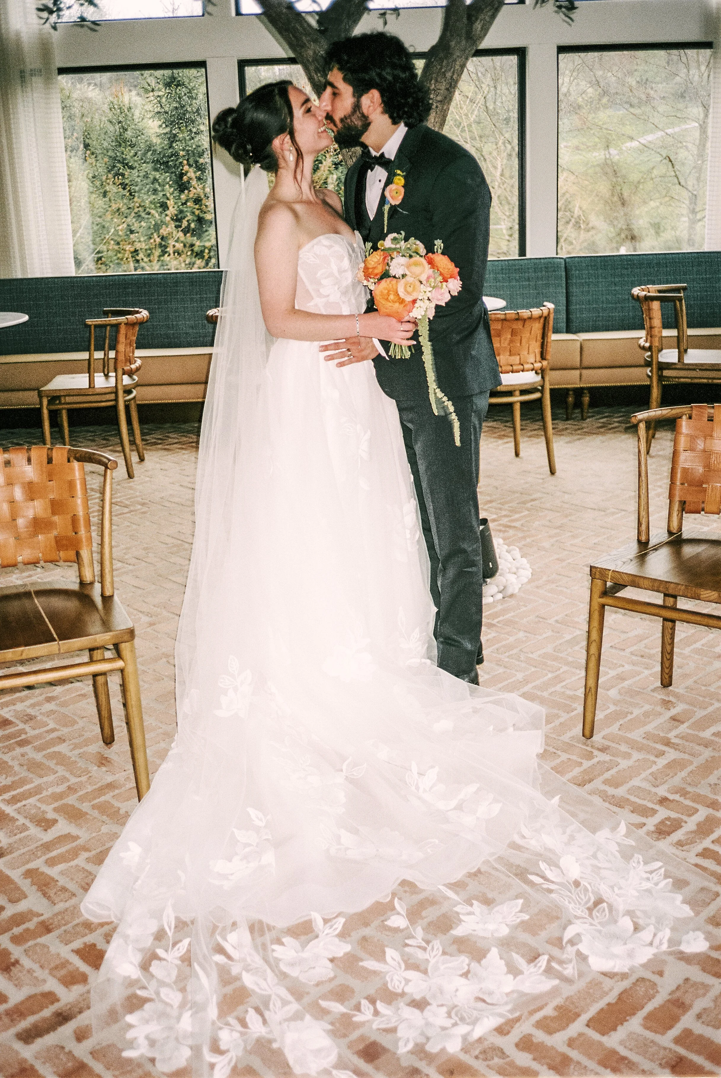 A bride and groom are standing close together indoors, touching noses, with the bride holding a bouquet of orange, pink, and white flowers. The bride is wearing a white strapless wedding gown with a long train and veil, and the groom is dressed in a black tuxedo with a bow tie and a flower boutonniere. Behind them, large windows reveal trees outside, and wooden chairs are arranged around the room.