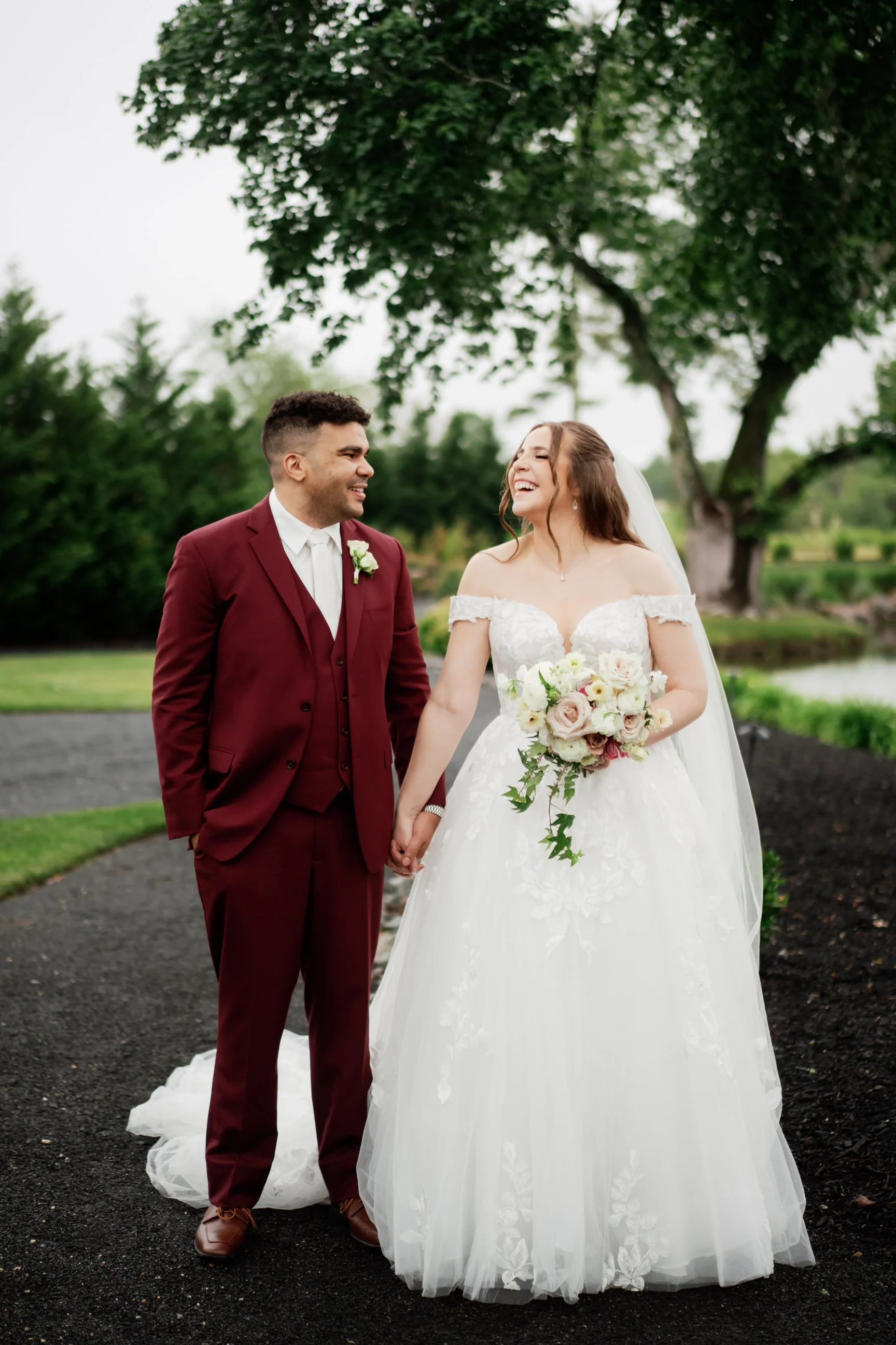 A smiling bride and groom holding hands outdoors on a cloudy day, standing on a dark gravel path with green trees and grass in the background. The bride is wearing a white princess-style wedding gown with off-the-shoulder lace sleeves, and holding a bouquet of white and blush roses. The groom is dressed in a burgundy suit with a white shirt, burgundy vest, and a white boutonniere.