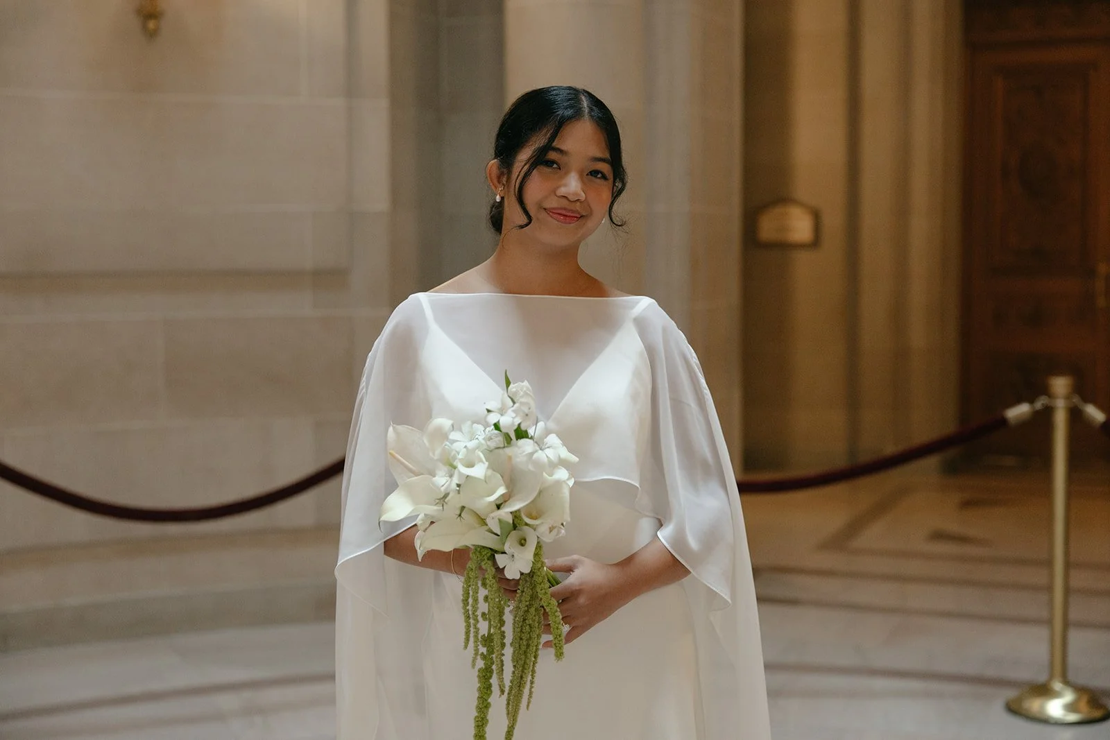 A woman in an elegant white dress holding a bouquet of white flowers, standing inside a building with stone walls and wooden doors.