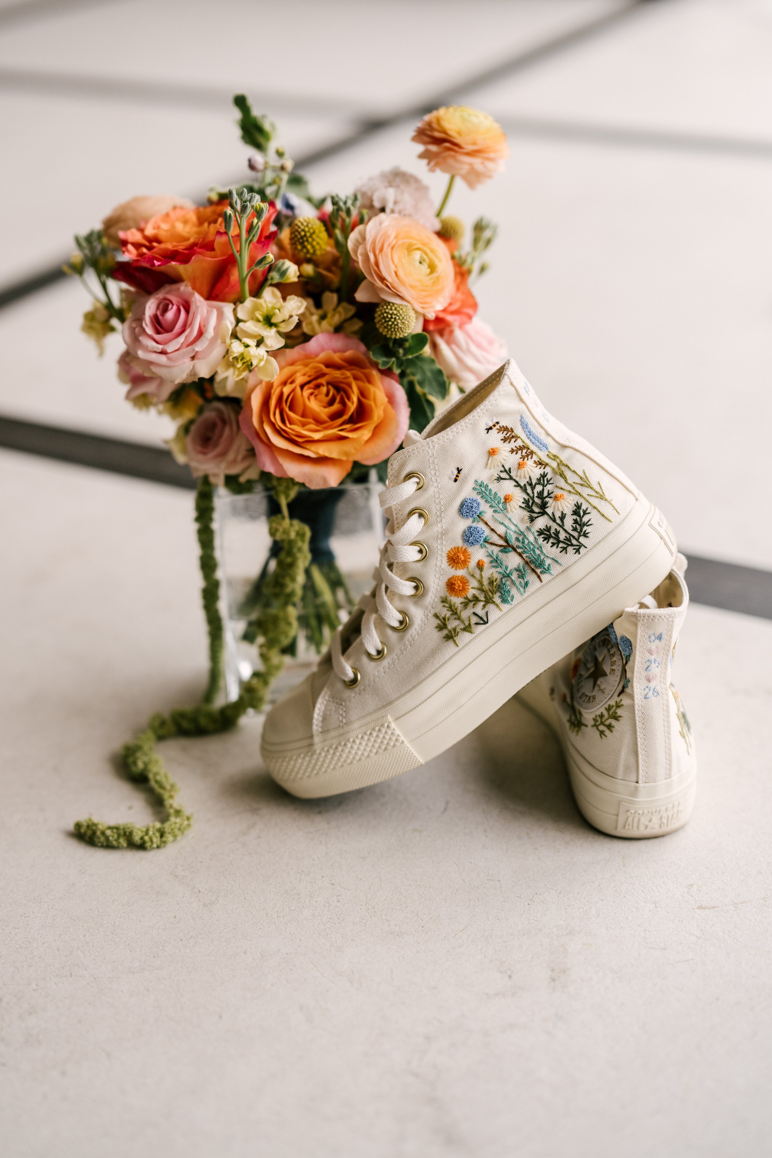 A pair of embroidered white canvas sneakers with floral designs leaning against a glass vase filled with a bouquet of assorted colorful flowers, including roses and ranunculus, on a white surface.