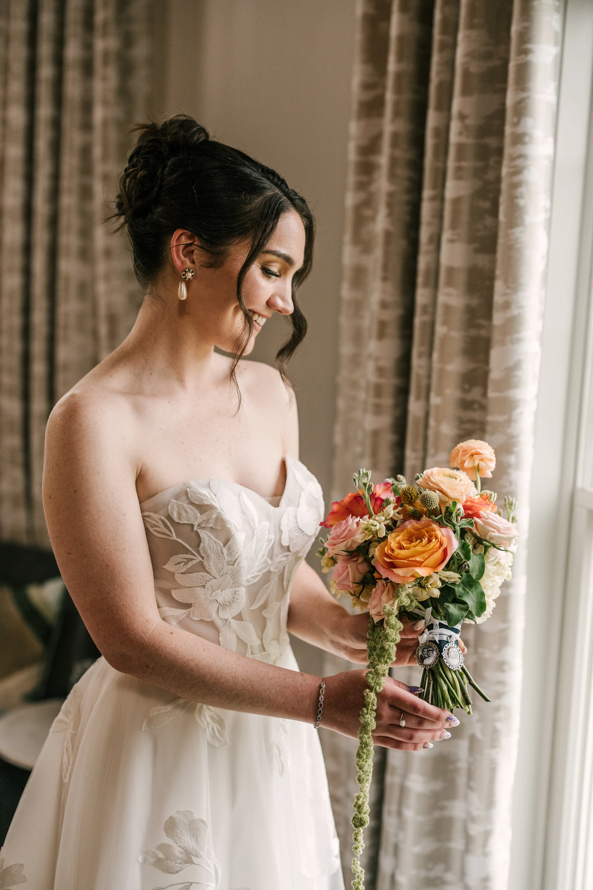 A woman in a wedding dress holding a bouquet of flowers near a window with curtains.