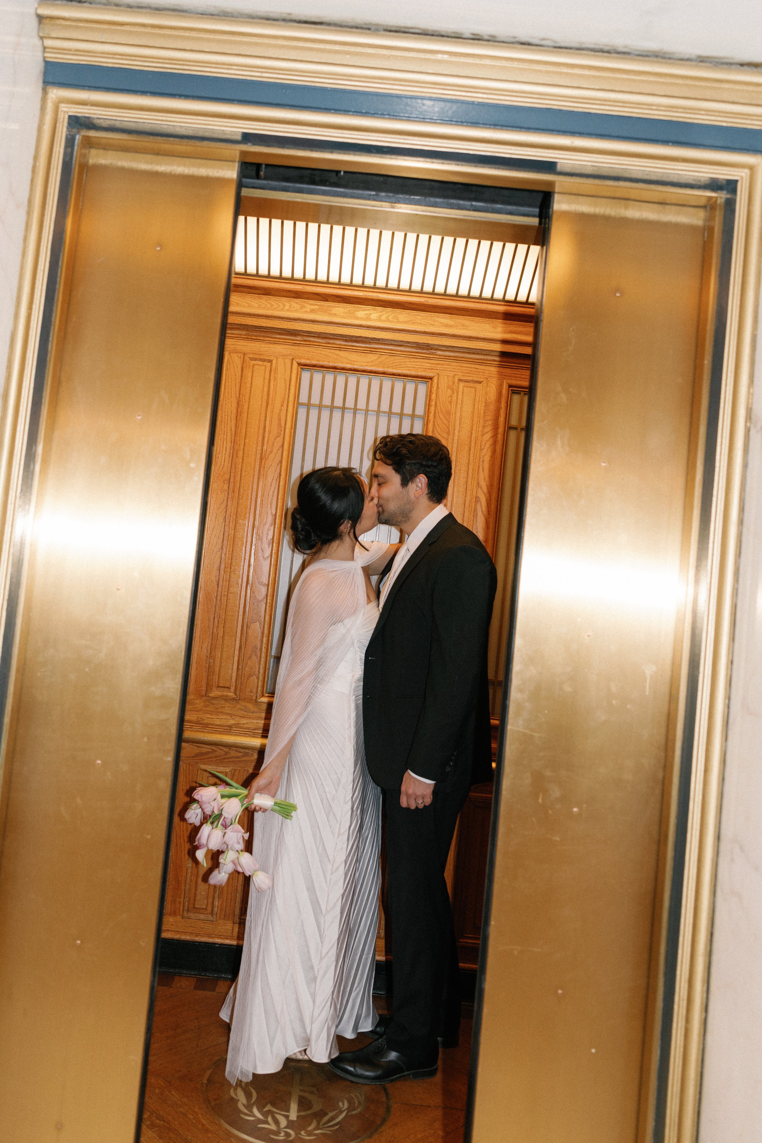 A couple in wedding attire kissing in front of a mirror, with the bride holding a bouquet of pink and white flowers.
