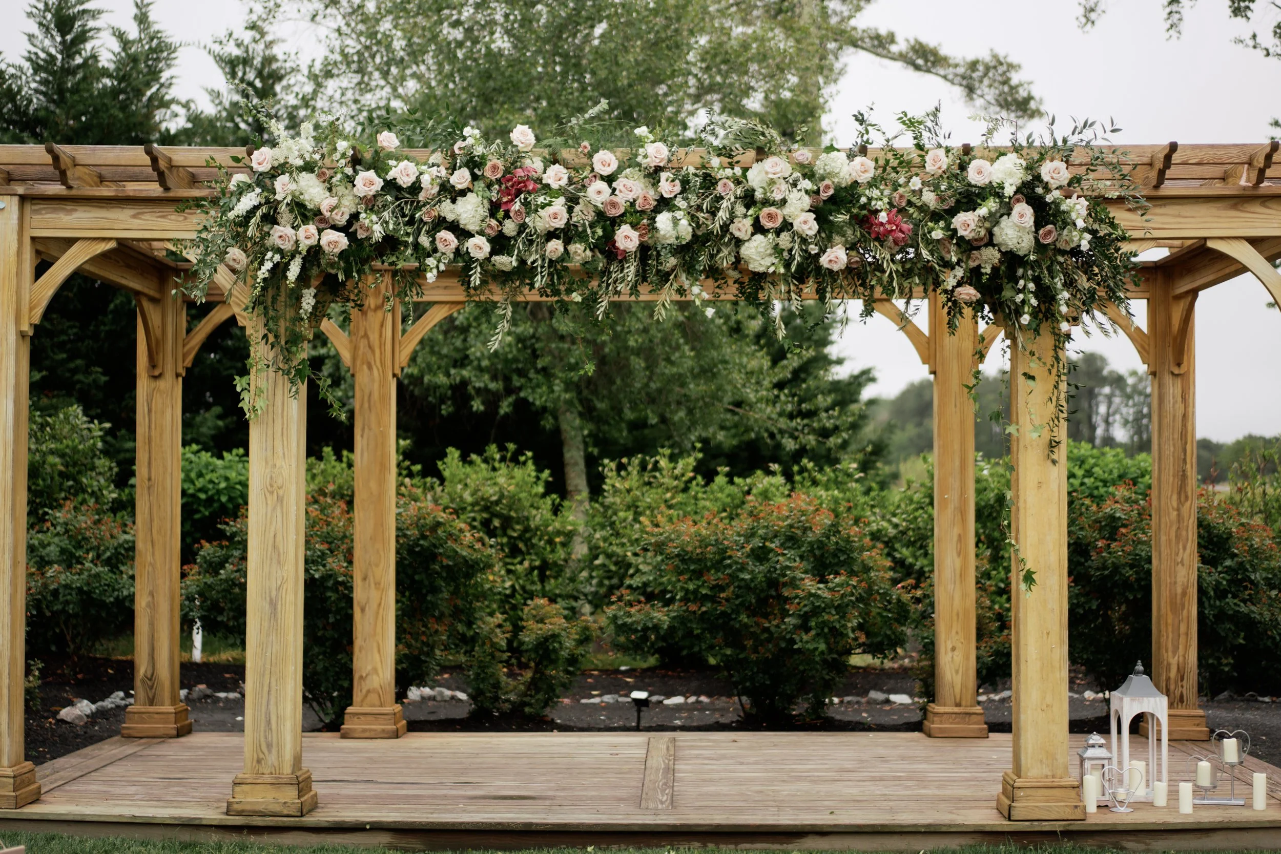 Wooden outdoor wedding arch decorated with white and blush pink flowers and greenery, set in a garden with trees in the background.
