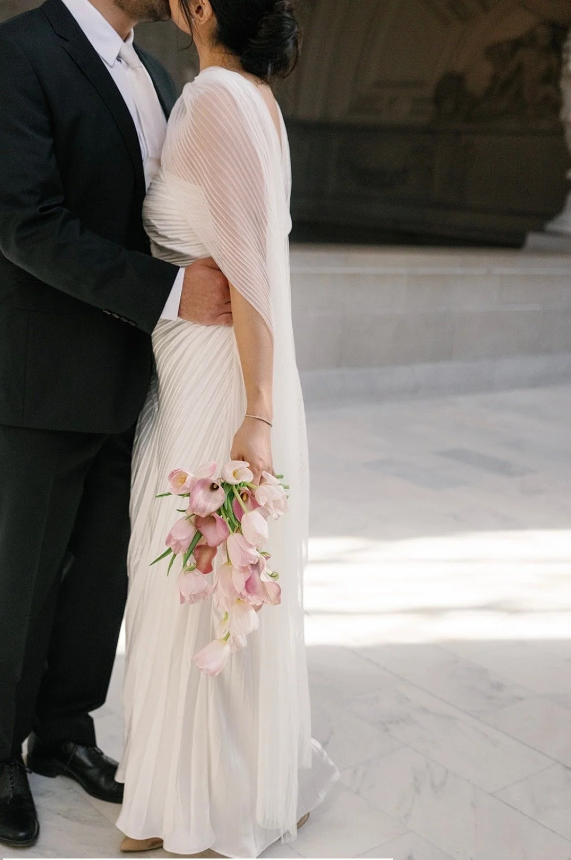 A bride in a white wedding dress holding a bouquet of pink and white calla lilies standing close to a groom in a black suit with a white shirt, in an indoor setting.