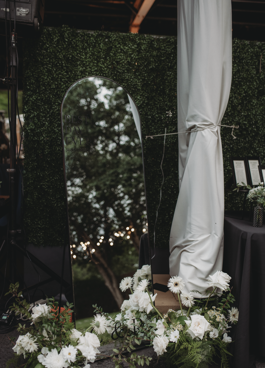 A reflective mirror with an inscription that reads "Best Day Ever" is surrounded by white flowers at a decorated event space, with greenery and a curtain in the background.