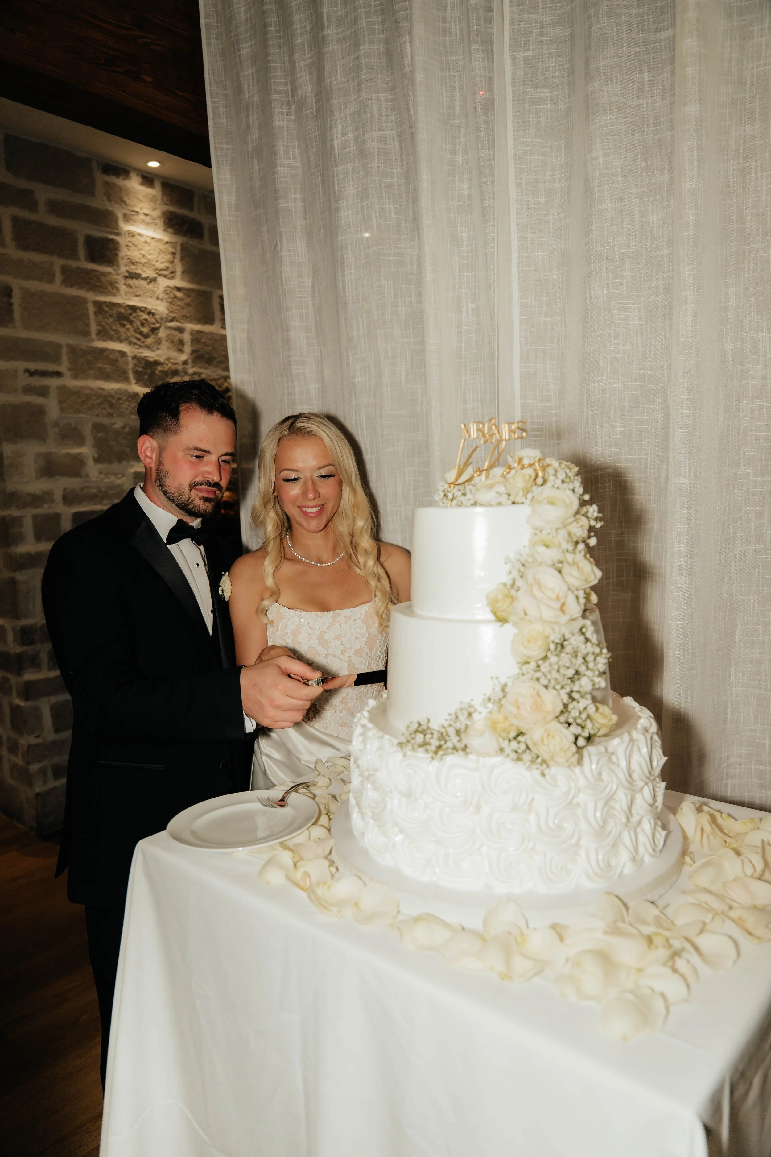 A newlywed couple, dressed in wedding attire, stands beside a three-tiered wedding cake decorated with white and cream-colored roses. The couple is smiling as they cut the cake together. The setting features a stone wall and light-colored curtains in the background.