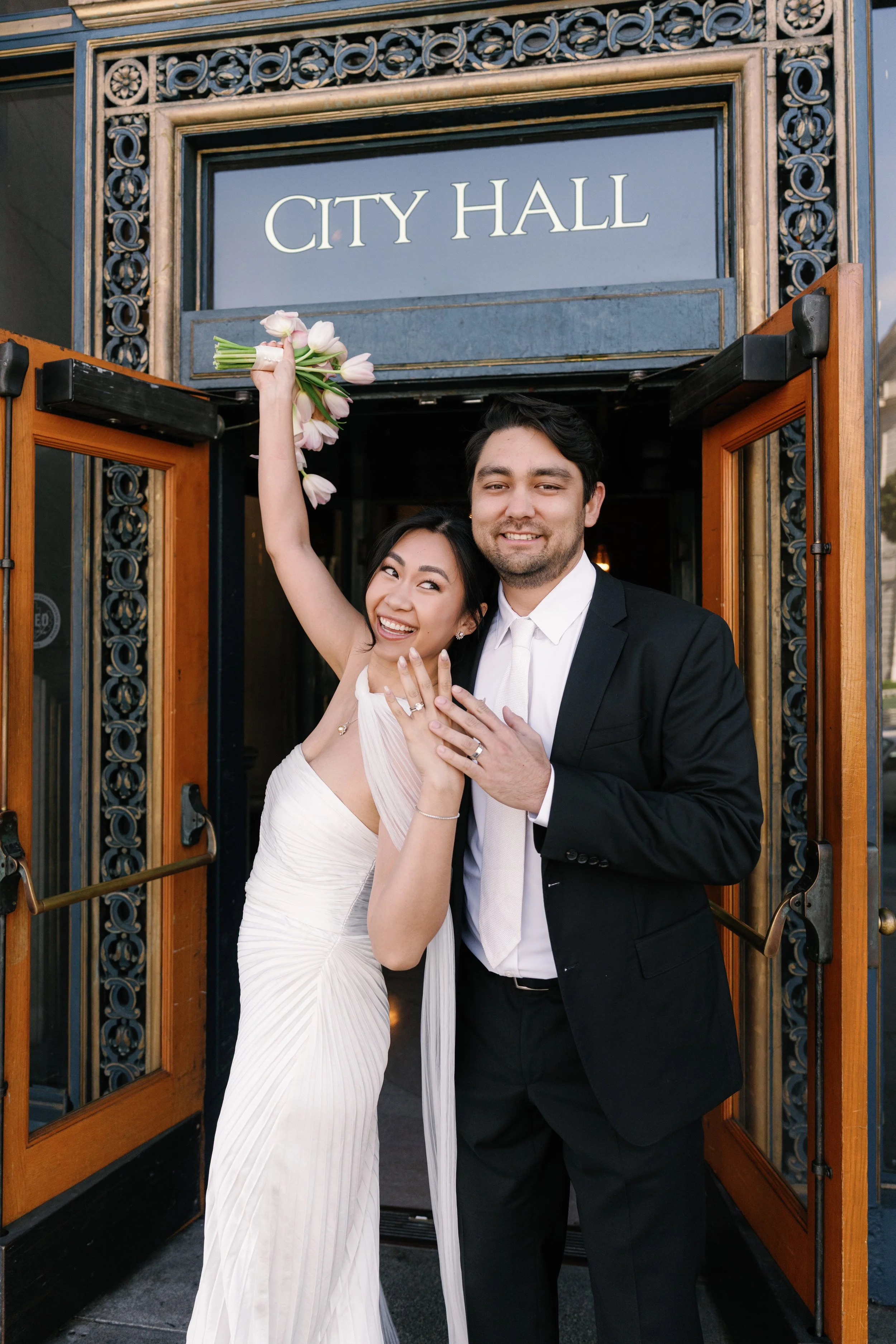 Two people, a woman in a white dress and a man in a black tuxedo, stand under a sign that reads 'City Hall.' The woman is holding pink tulips raised above her head, and they are smiling.