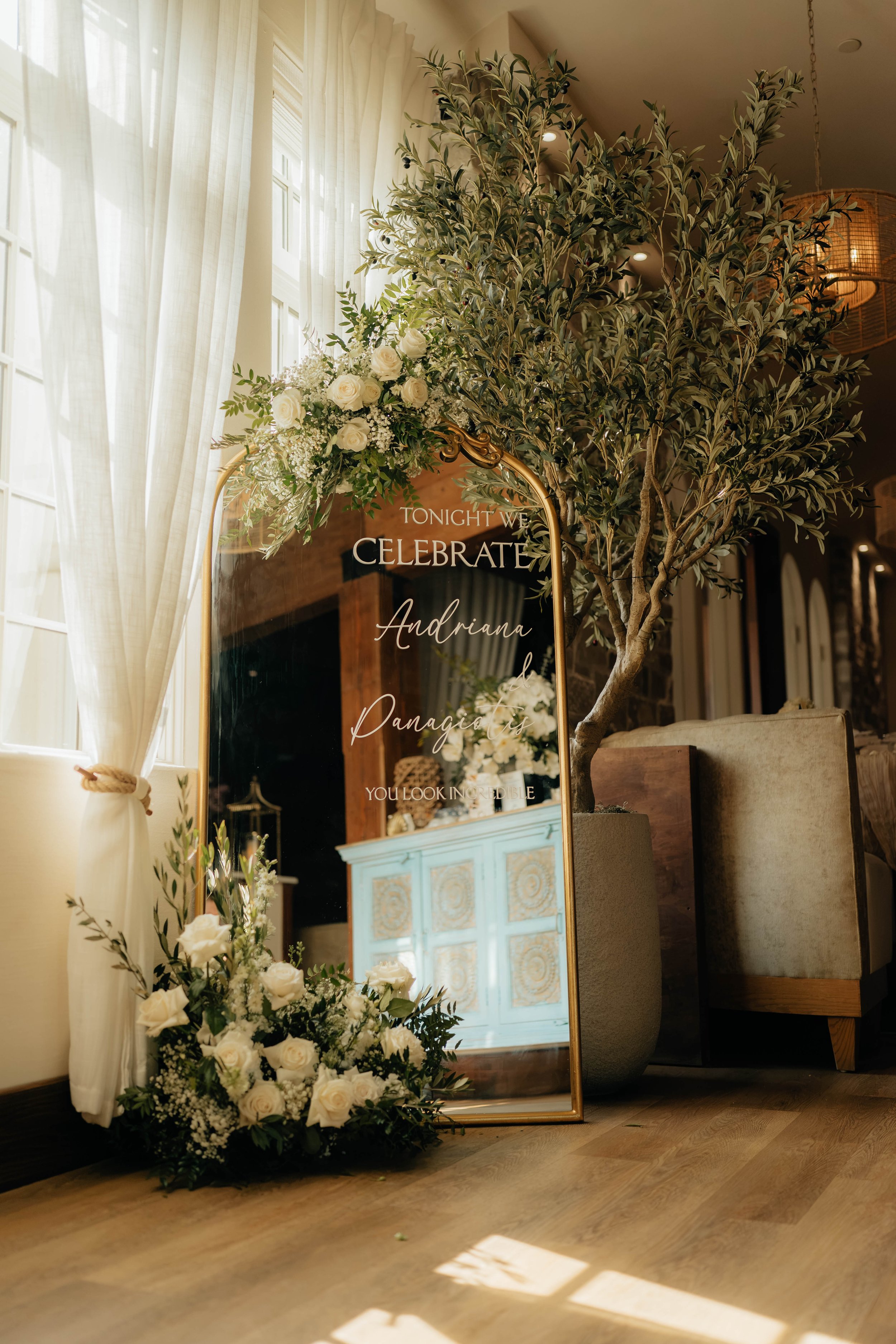 A decorative mirror with a floral arrangement of white roses and greenery on top, reflecting a wooden cabinet with ornate gold details inside a room with natural light coming through curtains.