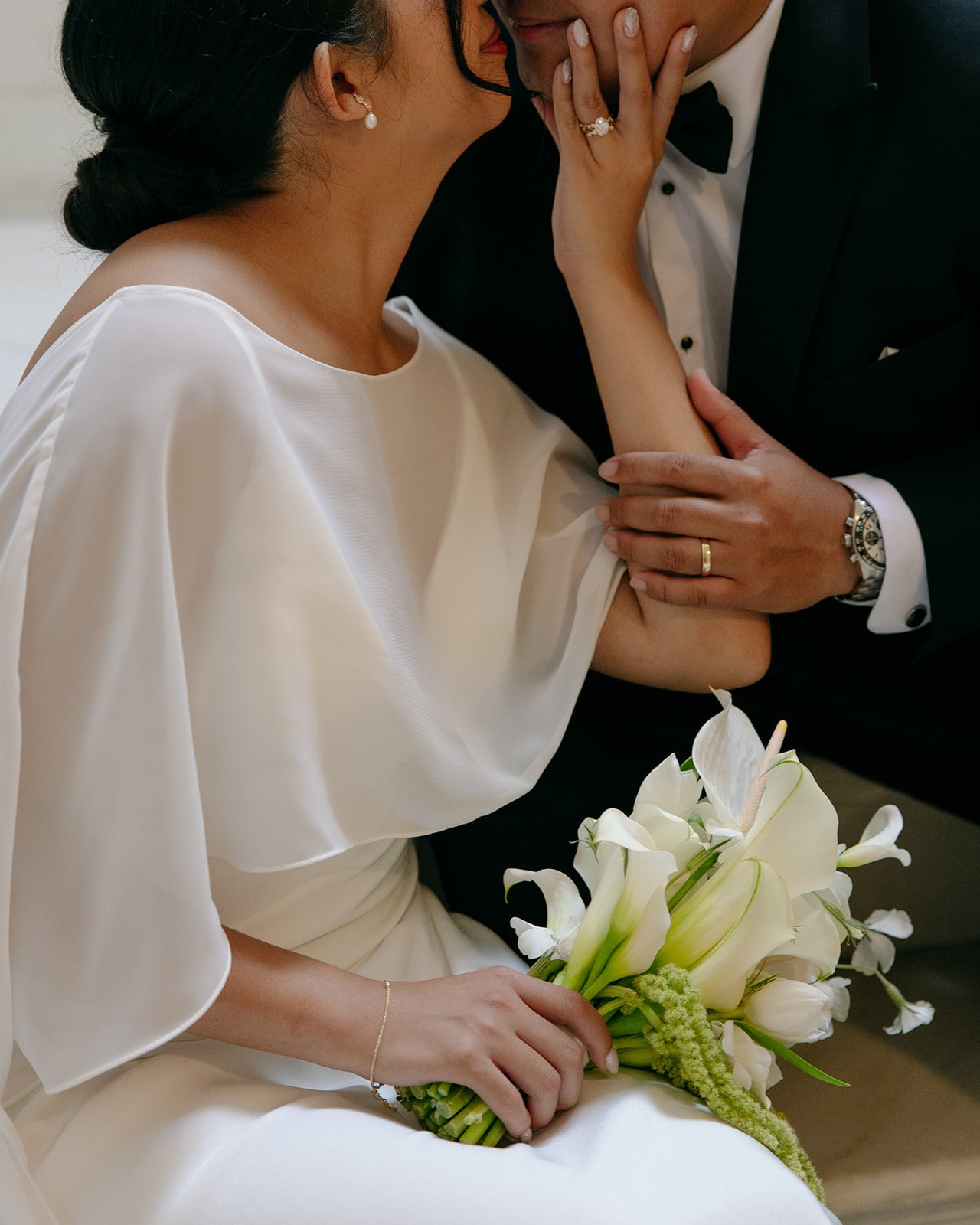 Close-up of a bride and groom during a wedding ceremony. The bride is wearing a white satin dress and pearl jewelry, holding a bouquet of white lilies. The groom is in a black tuxedo with a watch and wedding band, gently holding the bride's arm and touching her face.