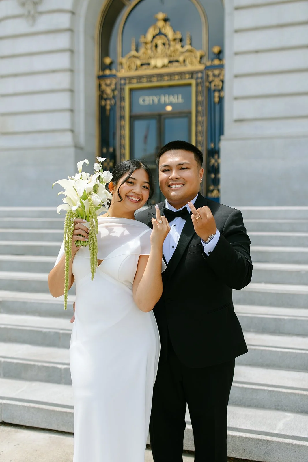 A newlywed couple in wedding attire standing on the steps in front of a city hall with ornate gold and blue decoration, smiling and showing their best wishes.