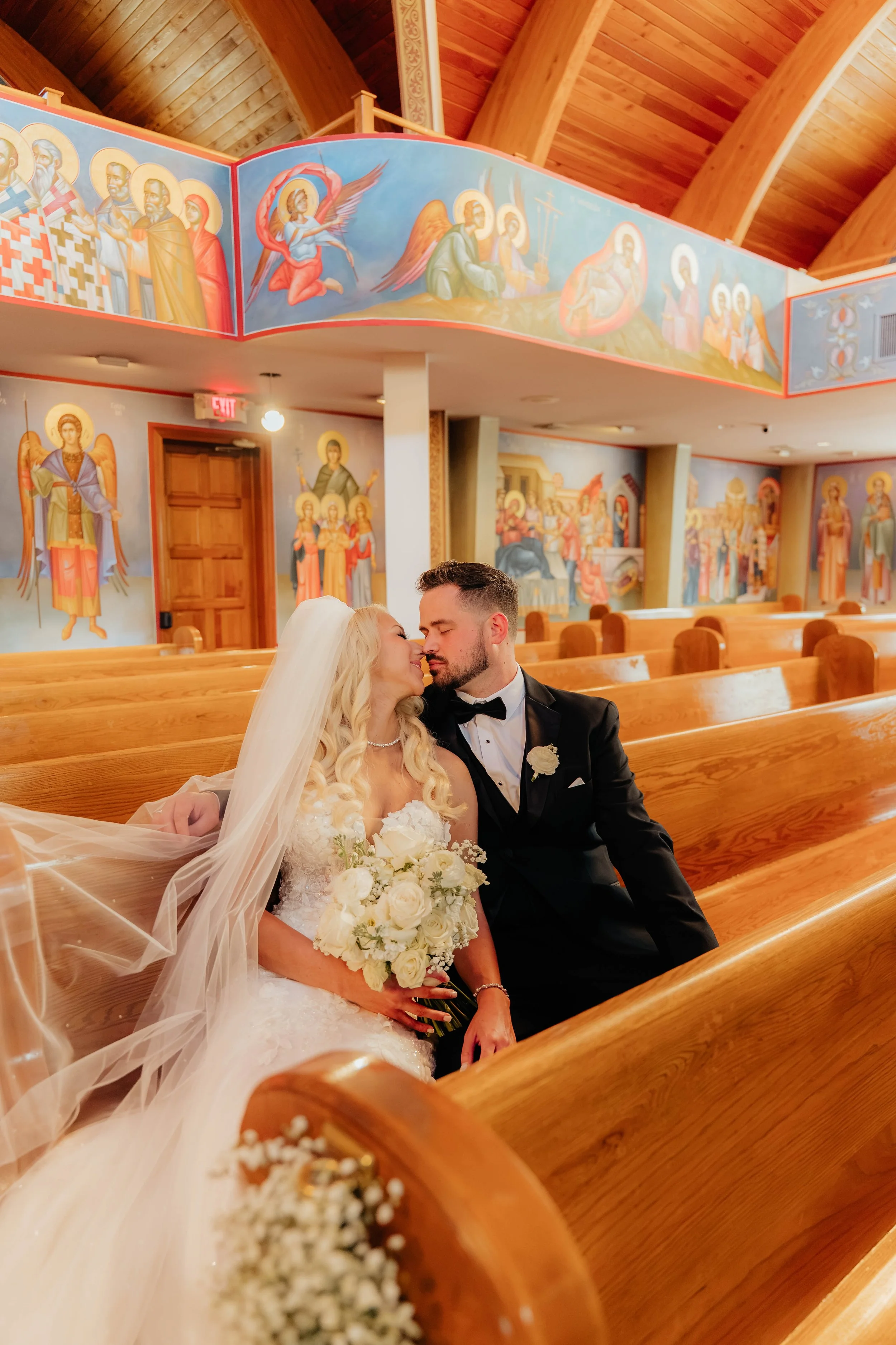 A bride and groom sitting closely in a church, smiling and leaning towards each other with their eyes closed. The bride has long blonde hair, a white veil, and is holding a bouquet of white roses. The groom is dressed in a black tuxedo with a bow tie. The church interior features wood pews, religious paintings on the walls, and a colorful mural on the balcony overhead.