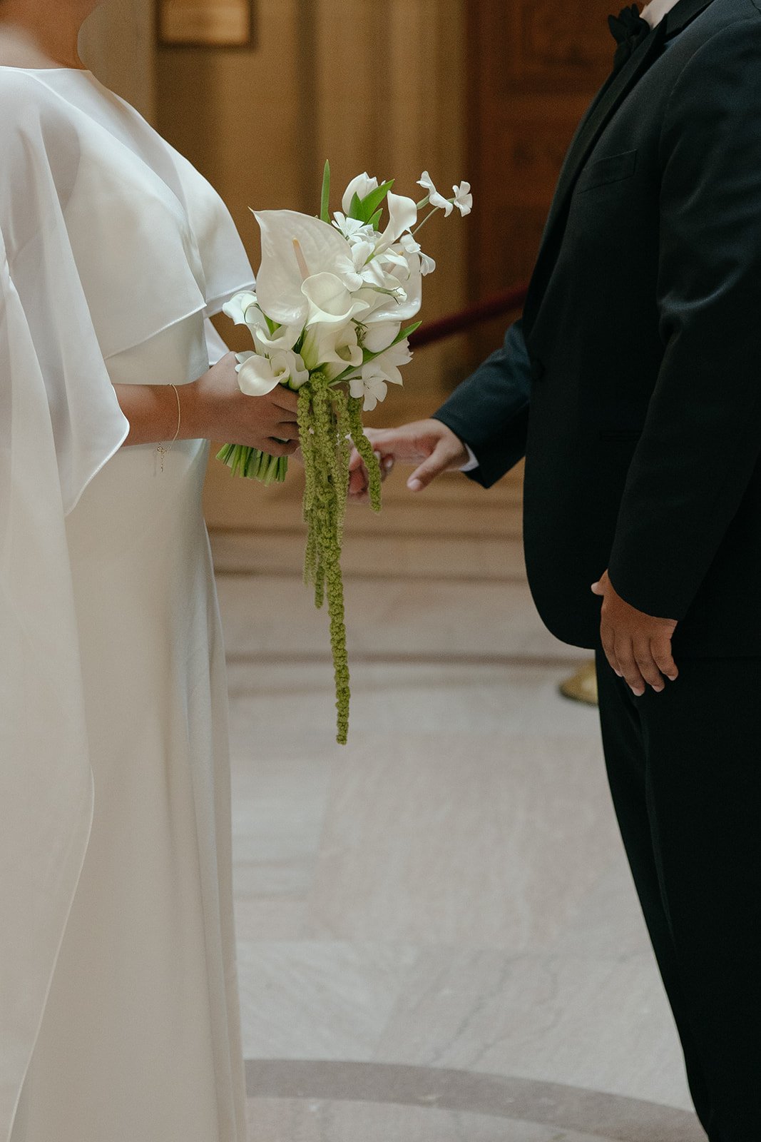 A bride holding a bouquet of white flowers, standing in front of a groom dressed in a black tuxedo and giving him a ring during a wedding ceremony.