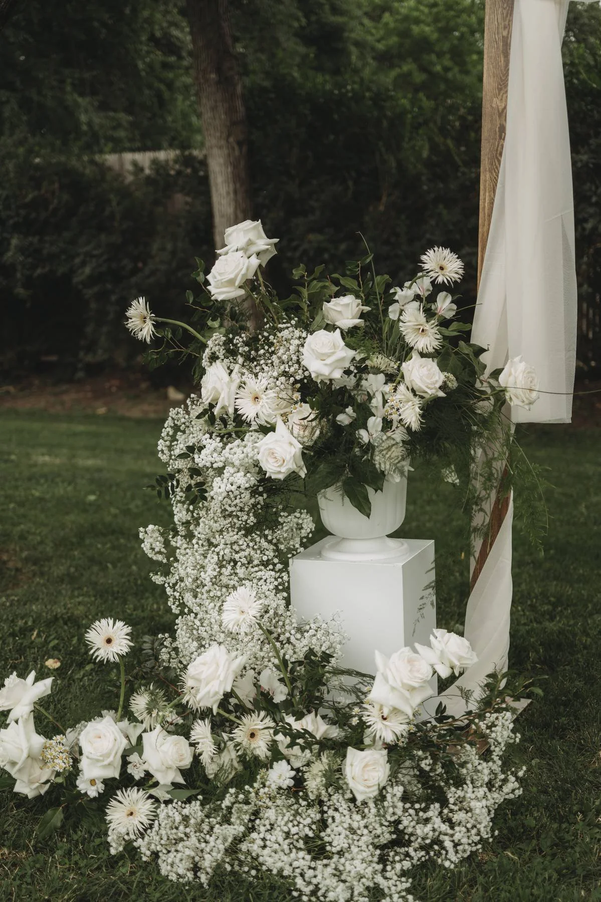 White floral arrangement with roses, daisies, baby's breath, and greenery on a white pedestal outdoors, near a wooden structure with white fabric draping.