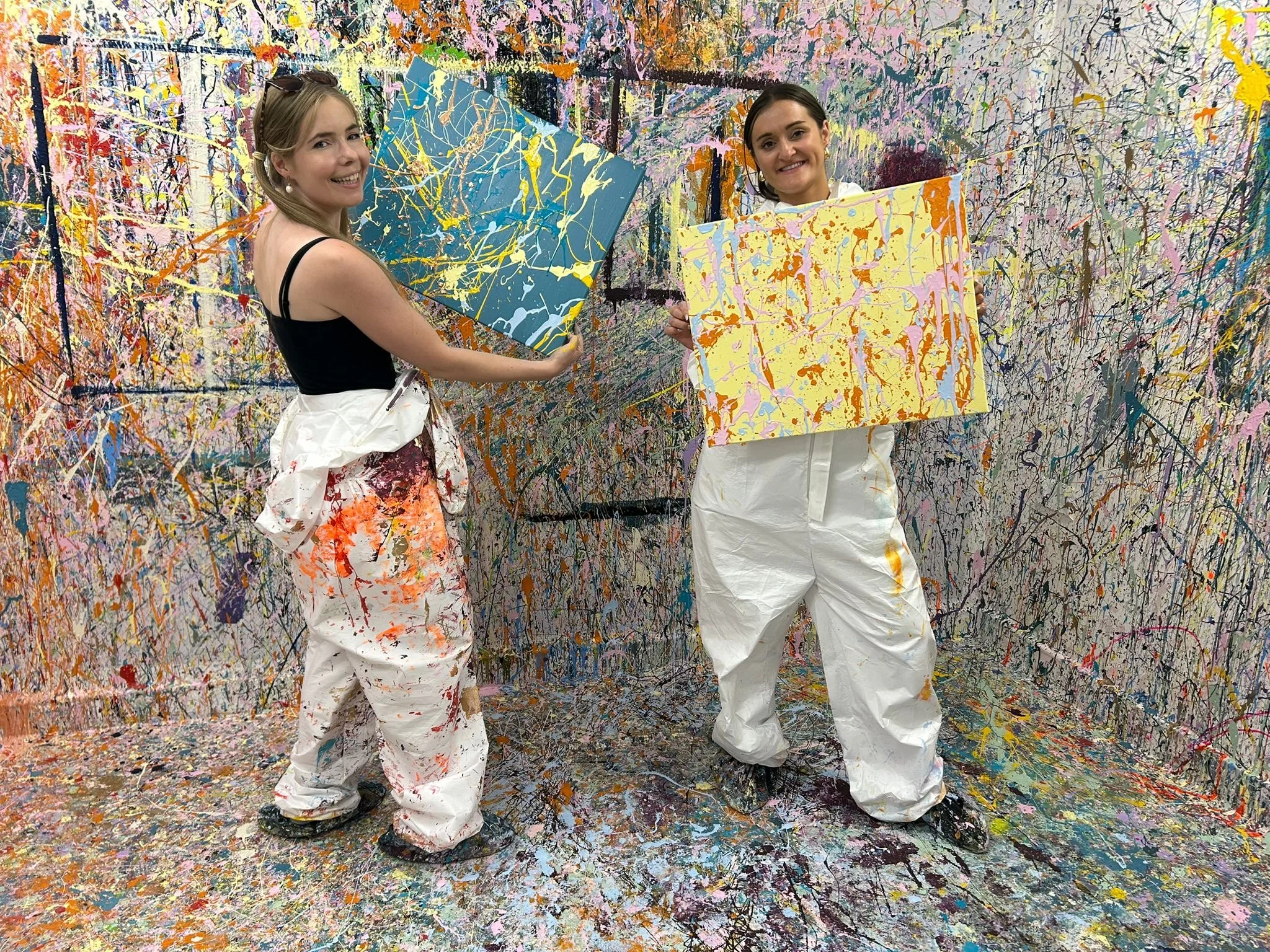 Two women in white splattered overalls holding up their abstract paintings in a colorful, splattered paint studio. The walls and floor are covered in multicolored paint splatters.