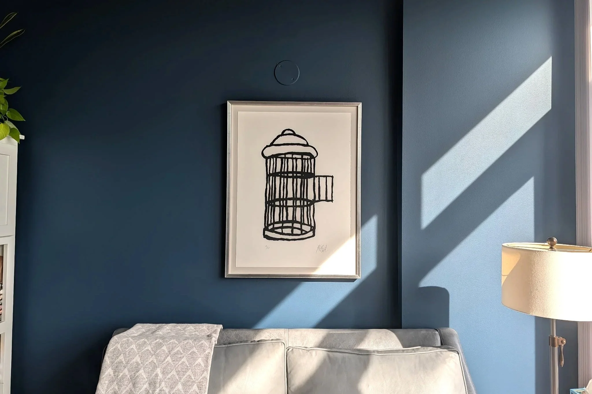 Photo of a dark blue painted wall with a light grey couch and black and white framed illustration of a birdcage.