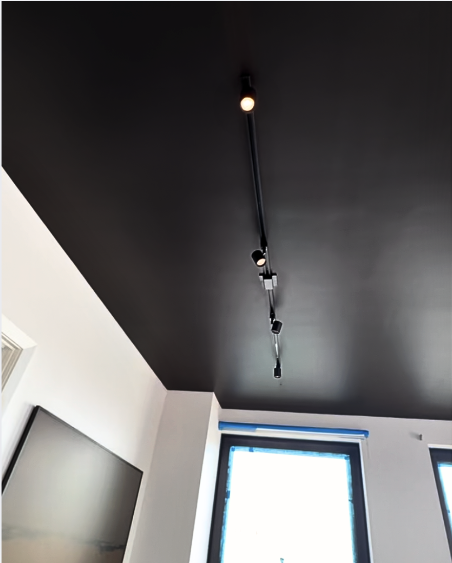 Dark brown ceiling with black track lighting and white walls. Blue painters tape is on the window frame which is being painted to match the dark ceiling paint.