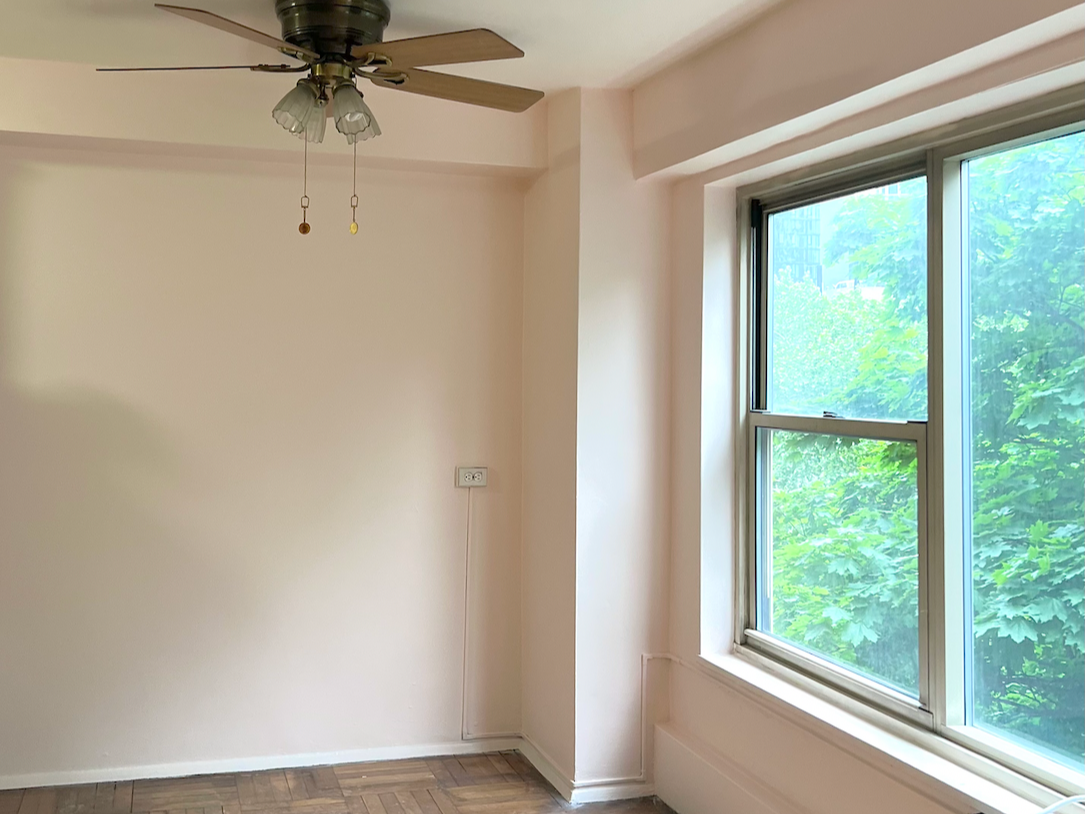 Photo of the corner and window of an unfurnished room in an apartment with walls freshly painted light pink and dark brown parquet flooring.