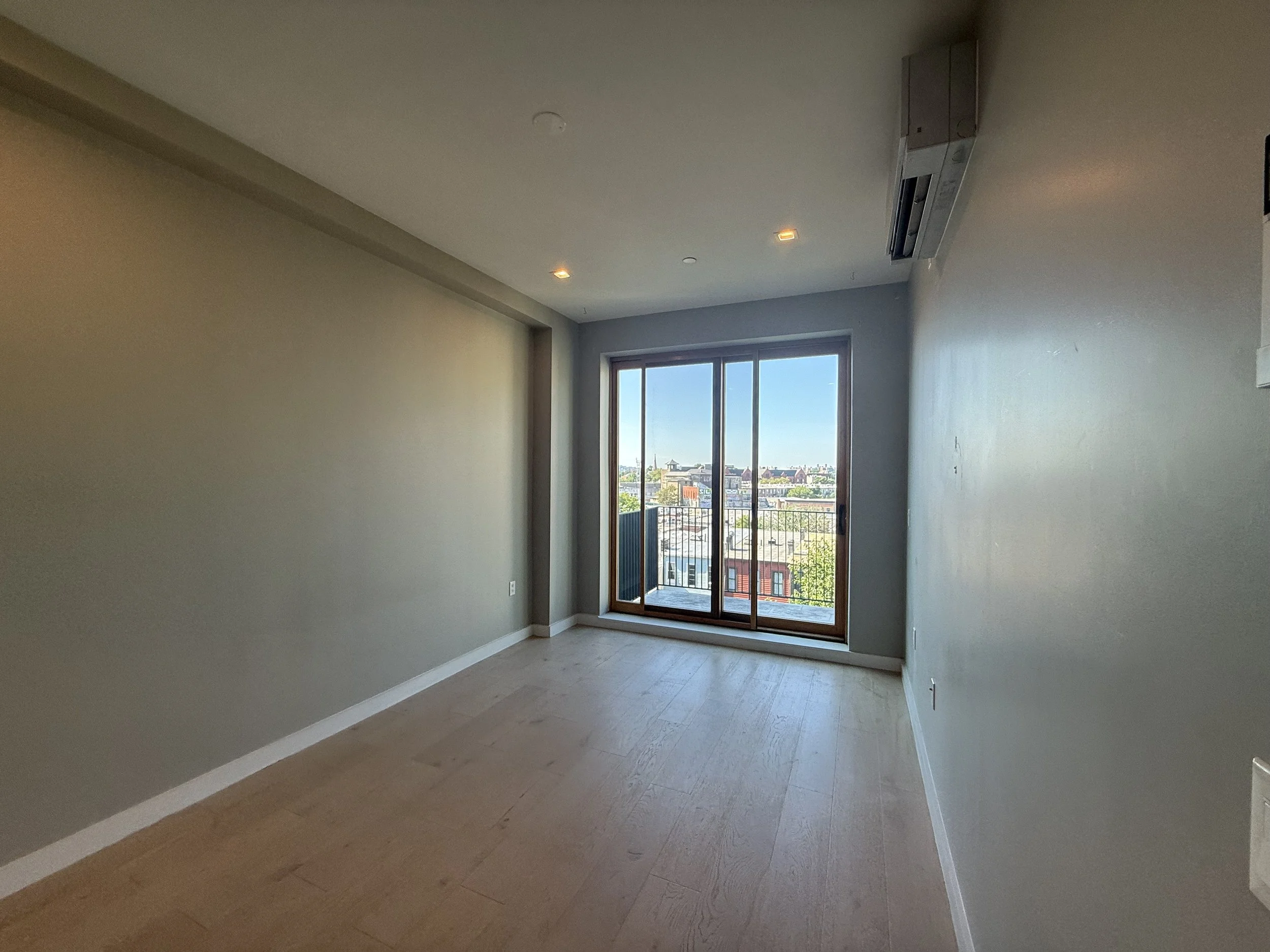 Before photo of an empty bedroom with wood floors, grey walls, and white trim with a floor to ceiling door leading out to a balcony
