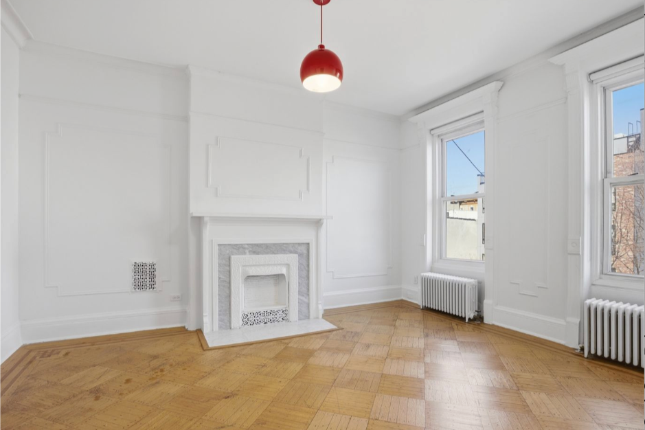 A bright white unfurnished bedroom with hardwood floors, fireplace mantle, high ceilings with molding detail, and a red chandelier light hanging from the ceiling.