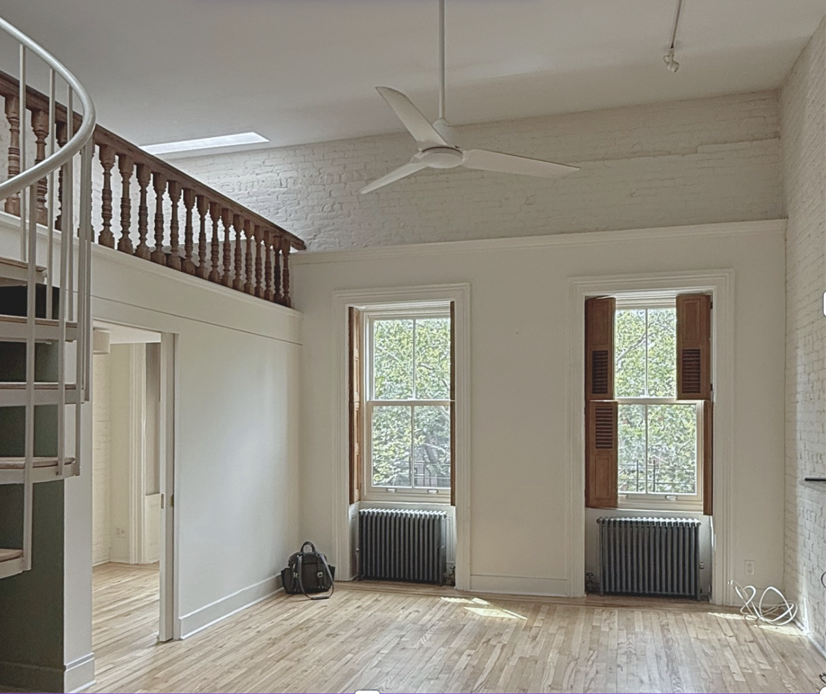 Interior photo of a brightly lit living room in a Brooklyn brownstone featuring white walls, 2 large windows and large spiral staircase off to the left.
