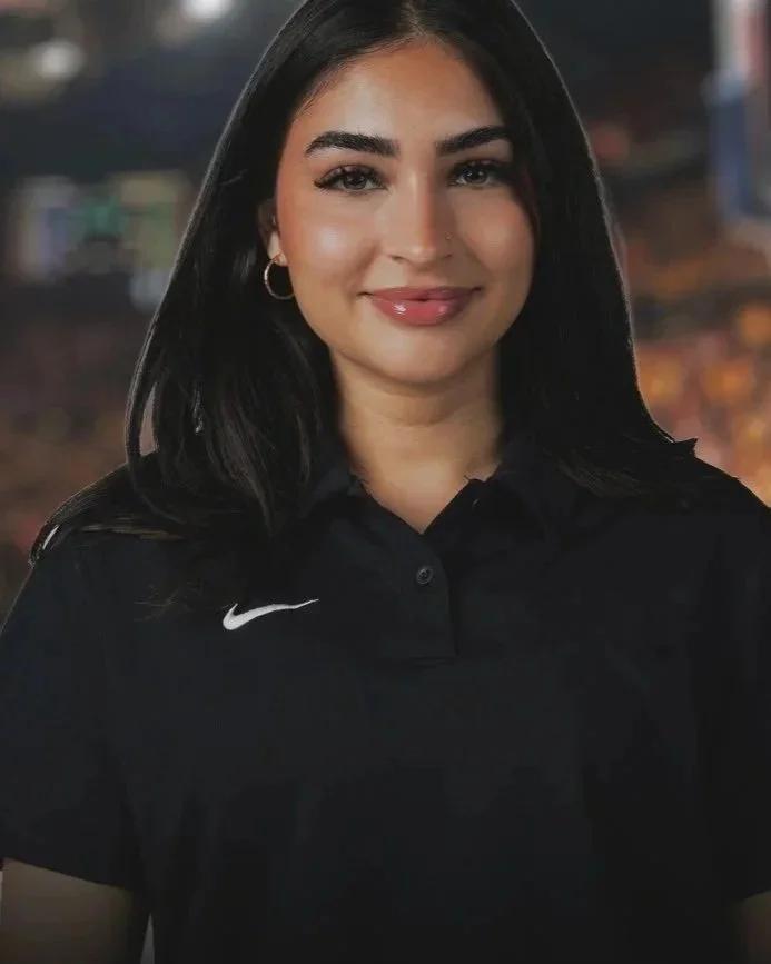 Young woman with dark hair and hoop earrings smiling, wearing a black Nike polo shirt, outdoors at dusk.