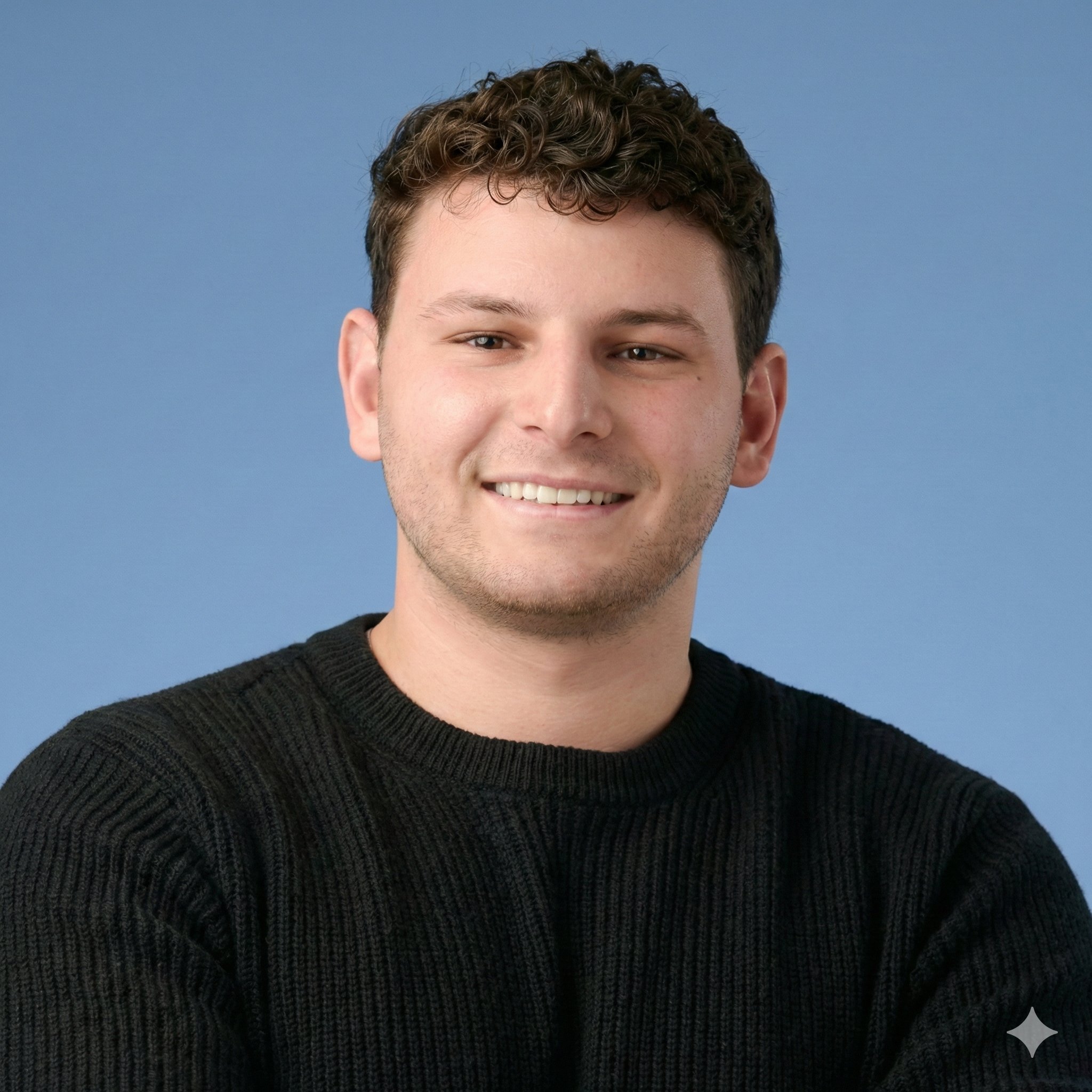 A young man with short curly brown hair smiling against a blue background, wearing a black sweater.
