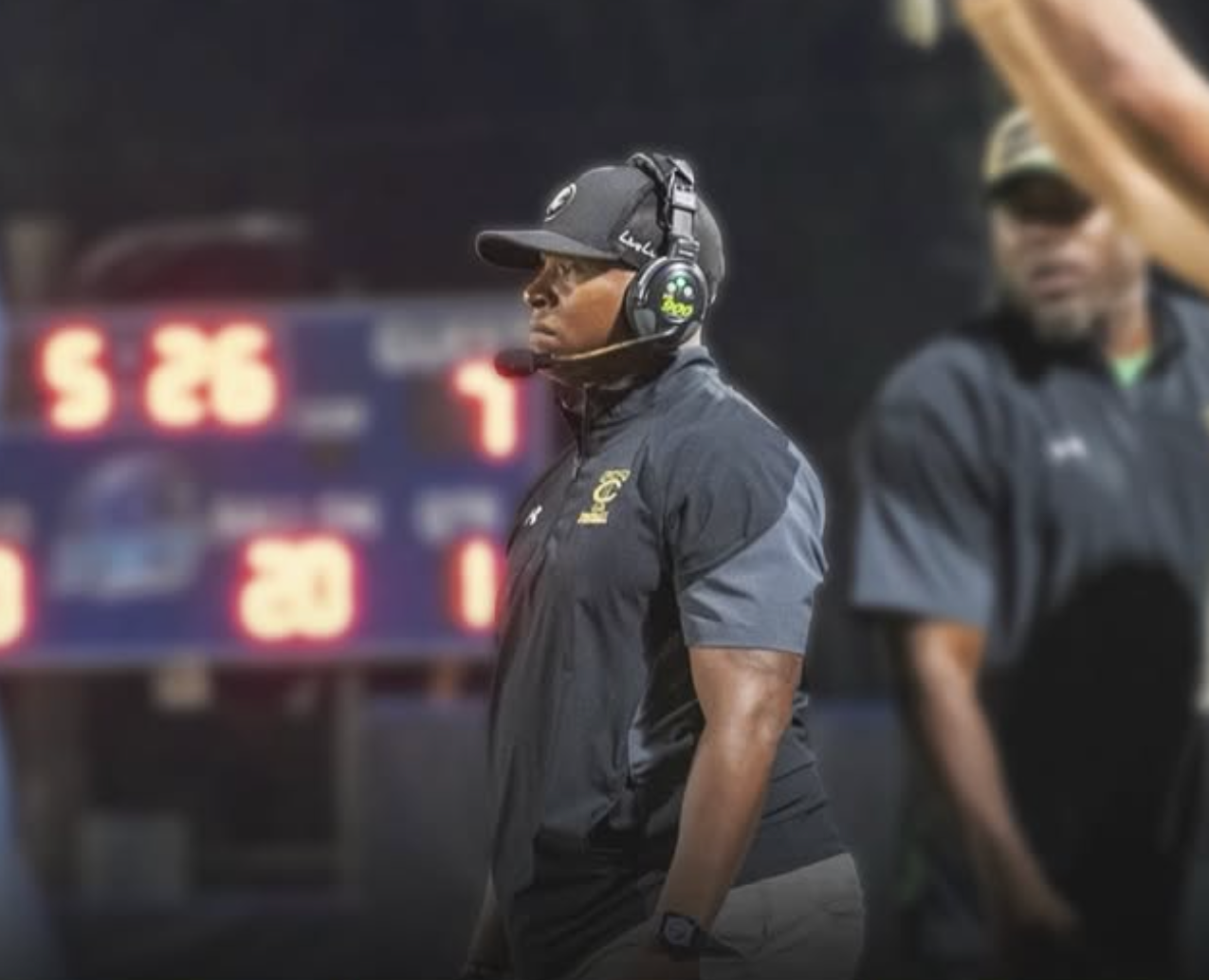 Two football coaches standing on the sideline during a game, with scoreboard in the background showing scores and time.