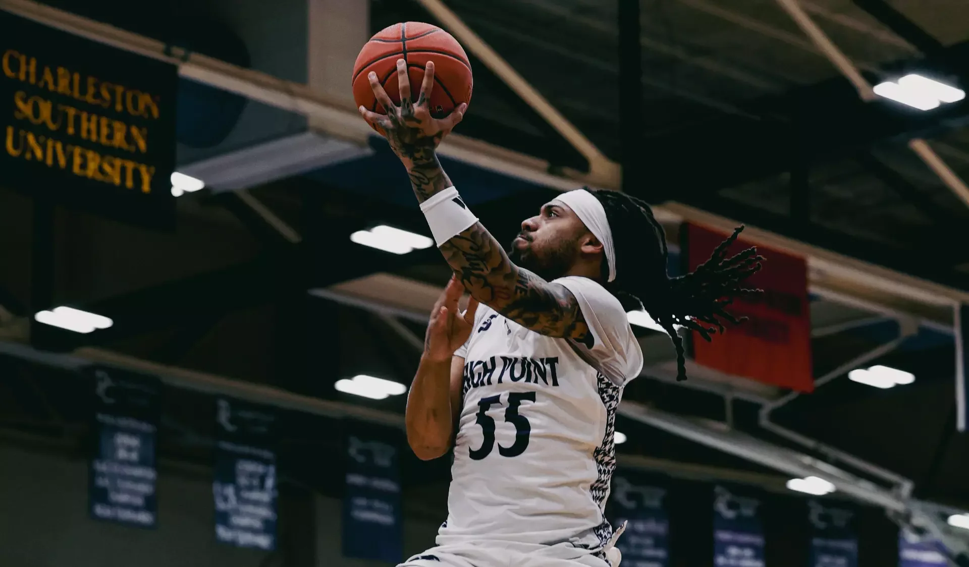 A basketball player with tattoos, wearing a white headband and jersey with the number 55, is jumping to make a shot in a gymnasium. Banners hang in the background, including one that reads 'CHARLESTON SOUTHERN UNIVERSITY'.