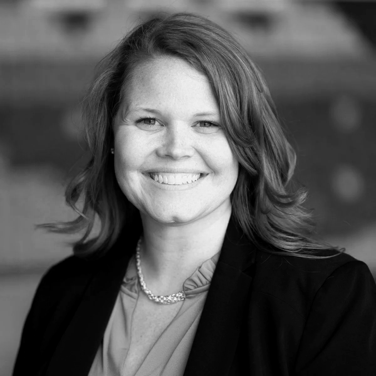 Black and white portrait photo of a smiling woman with shoulder-length hair, wearing a blazer, blouse, and a necklace.