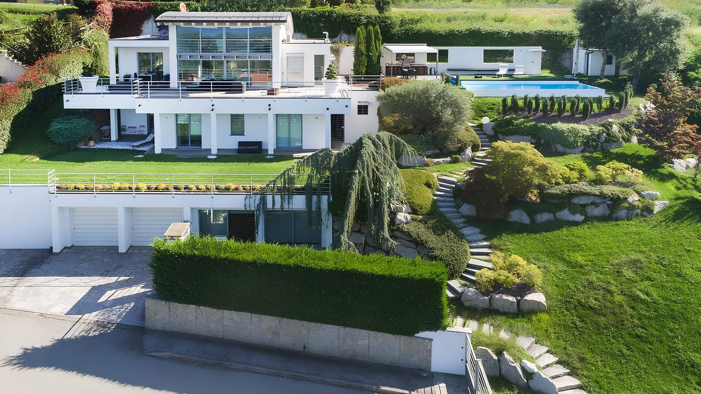 Modern multi-level house with large glass windows, multiple outdoor terraces, a swimming pool, landscaped garden with trees and bushes, and stone pathways.