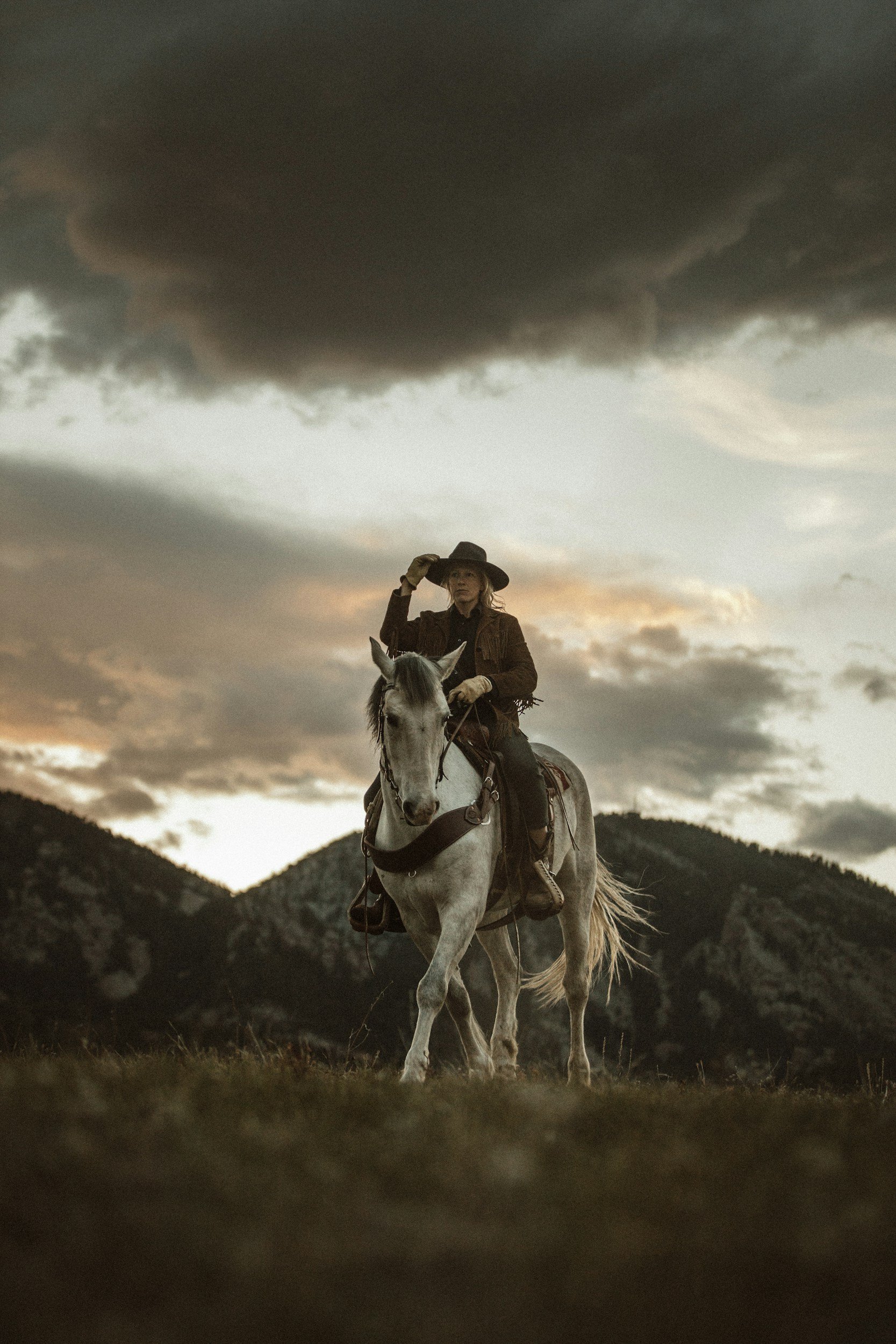A woman riding a white horse in a mountainous landscape during sunset with dark clouds overhead.