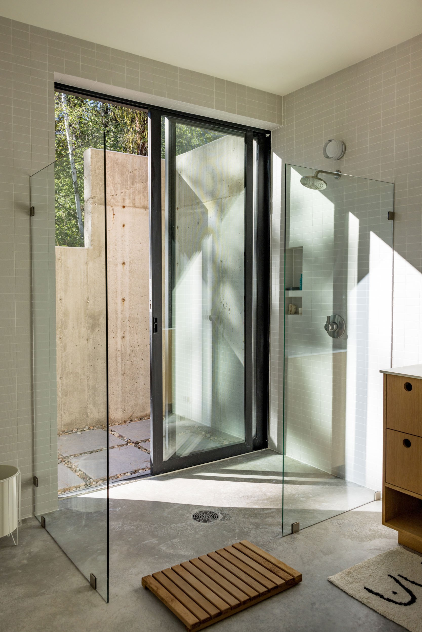 Modern bathroom with glass shower doors, an outdoor view through a large window, a wooden mat on the floor, and light wood cabinetry.