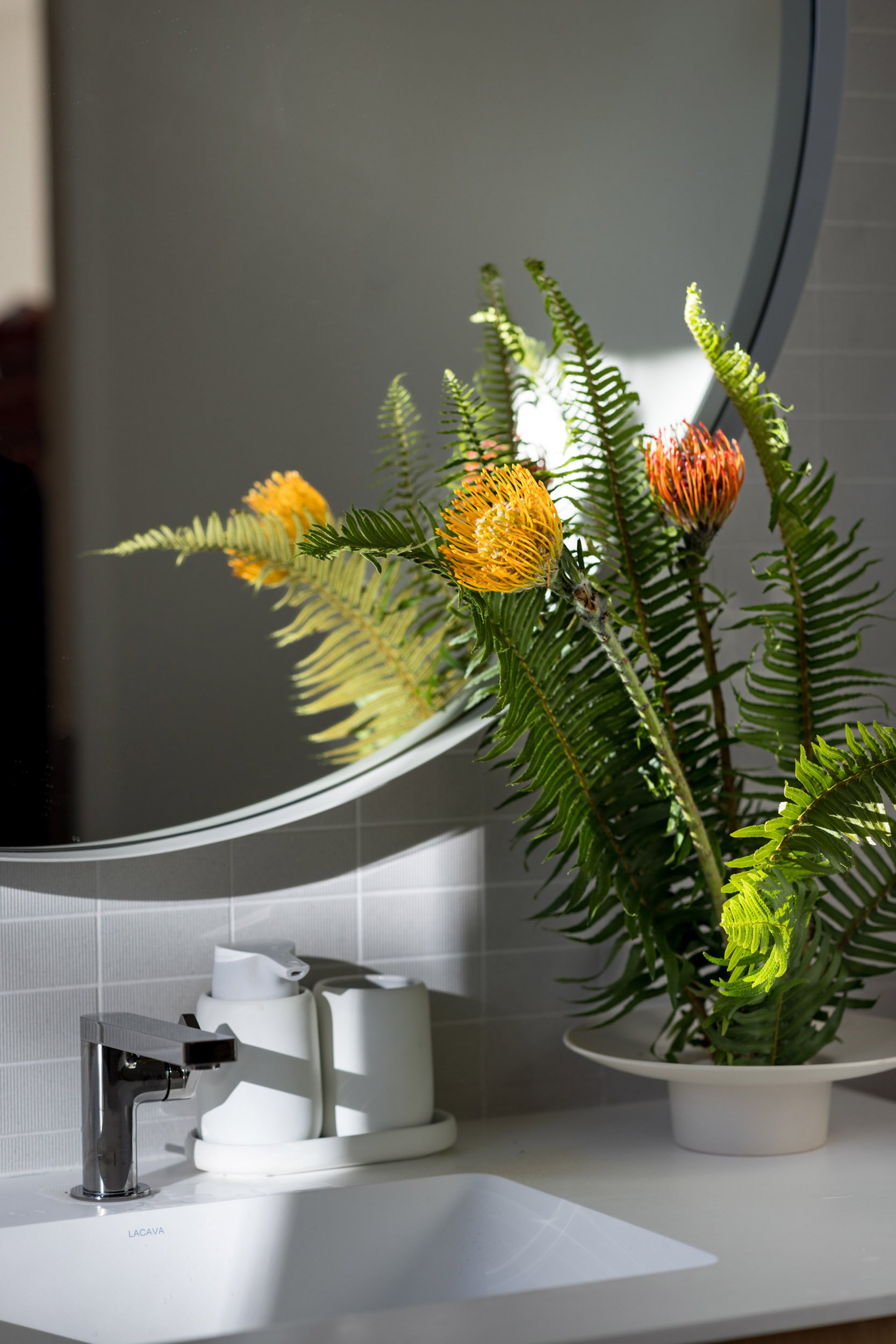 Bathroom sink area with faucet, soap dispenser, and a vase with vibrant protea flowers and ferns, reflected in a round mirror.