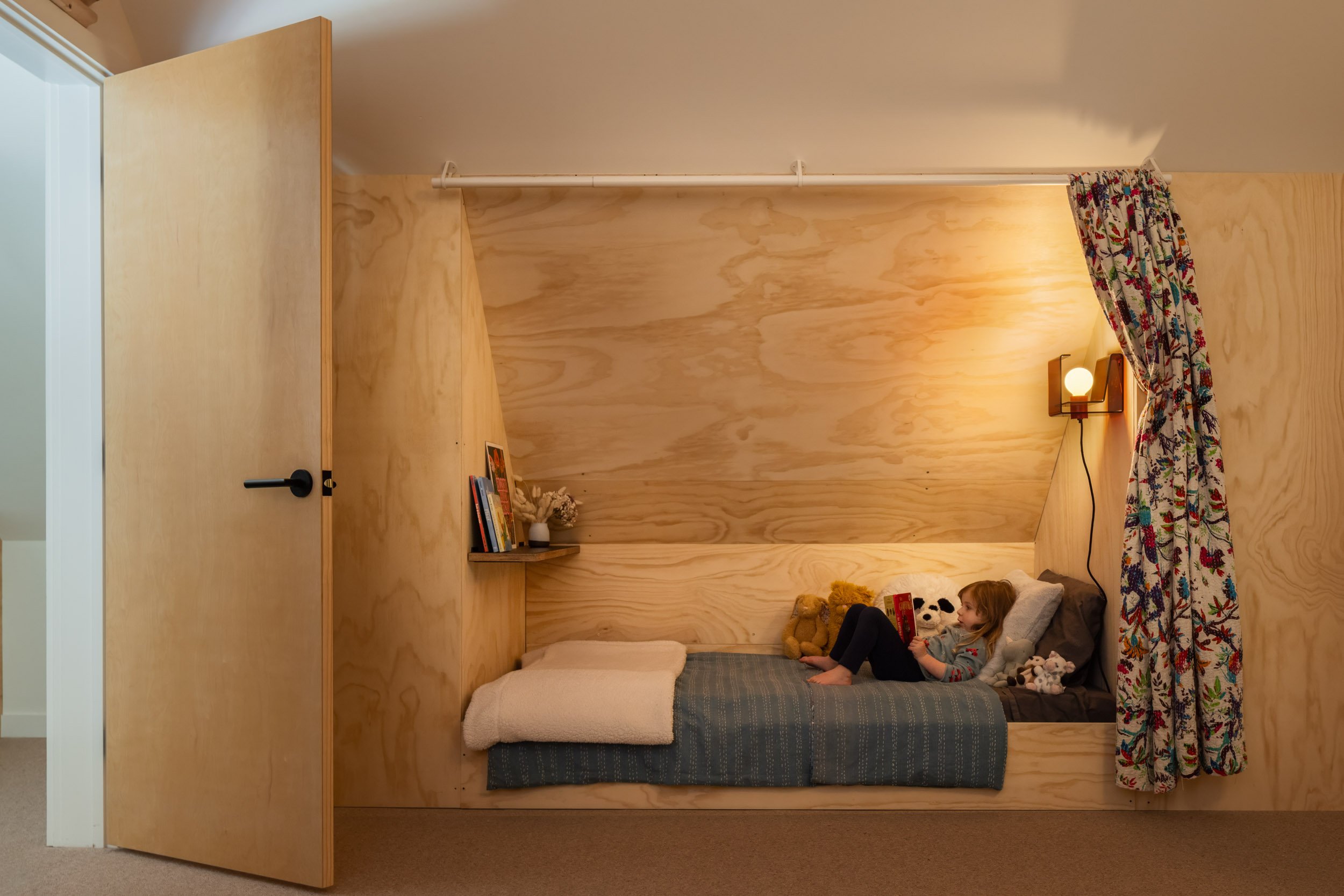 Child reading in a cozy wooden alcove bed with toys, a floral curtain, and a wall-mounted lamp.