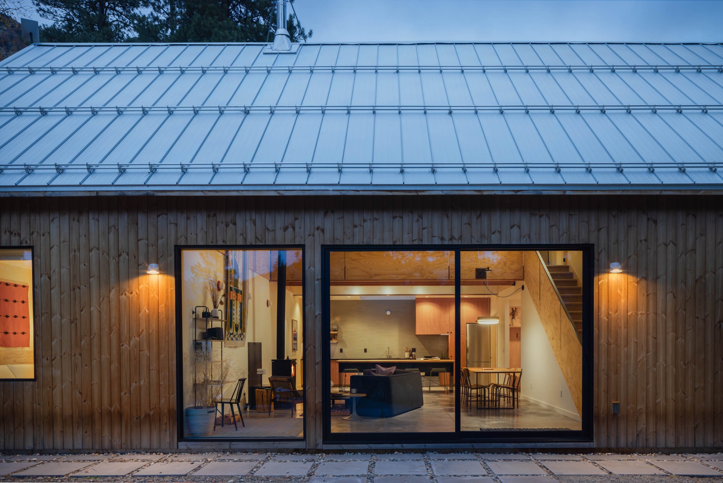 Exterior view of a modern house with wooden siding and large windows revealing the interior. The house has a metal roof and outdoor lighting on either side of the windows. Inside, the kitchen and living area are visible, featuring modern furnishings