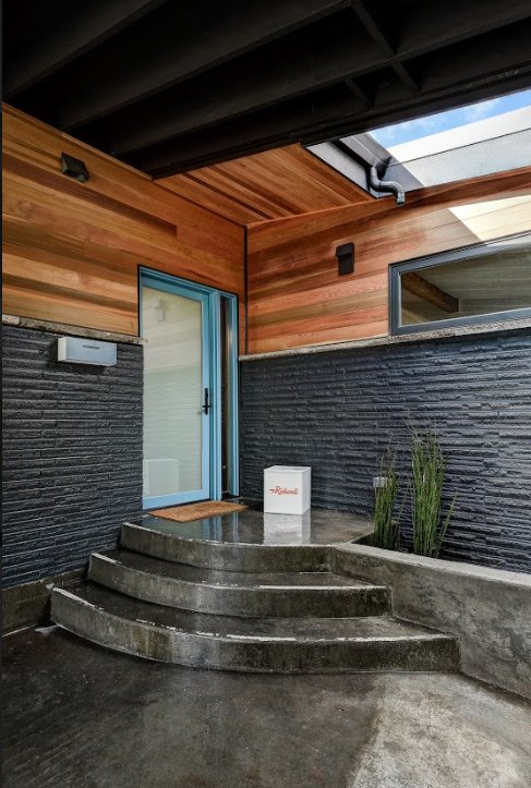 Modern house entrance with blue-framed glass door, curved concrete steps, wood paneling, and a small rug. Brick wall with plants and package at the door.