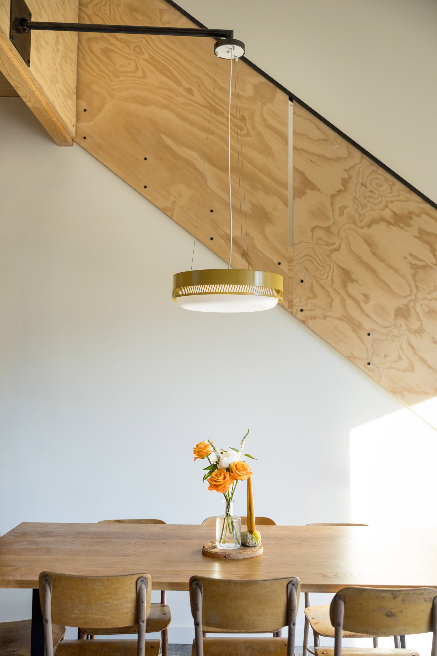Dining room with wooden table and chairs, a vase with orange flowers, and a modern hanging light fixture, in front of a sloped wooden wall.