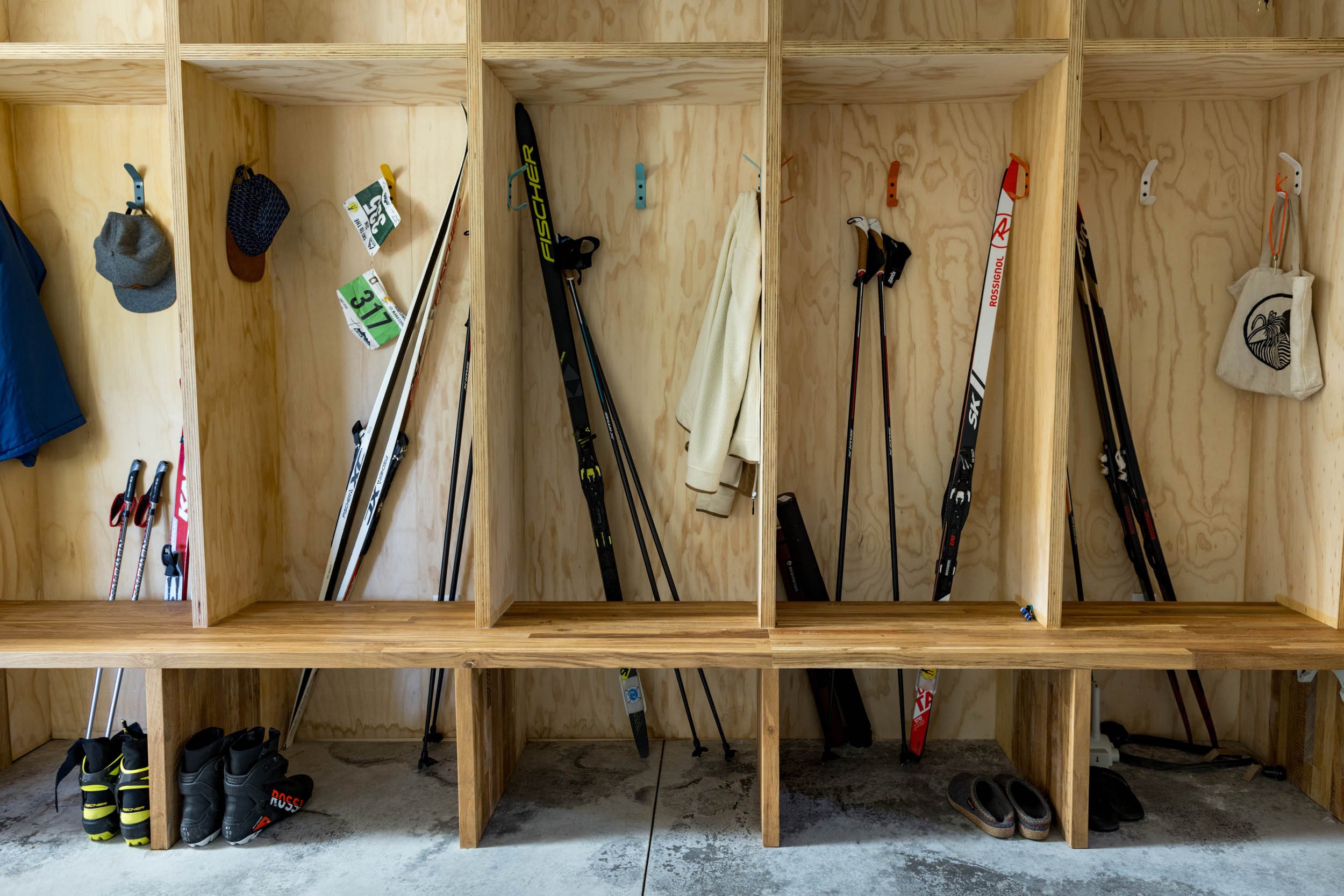 A wooden storage area with cubbies containing ski equipment, including skis, ski poles, and boots. Some hats and jackets are hanging on hooks, along with a small tote bag. The floor is concrete.