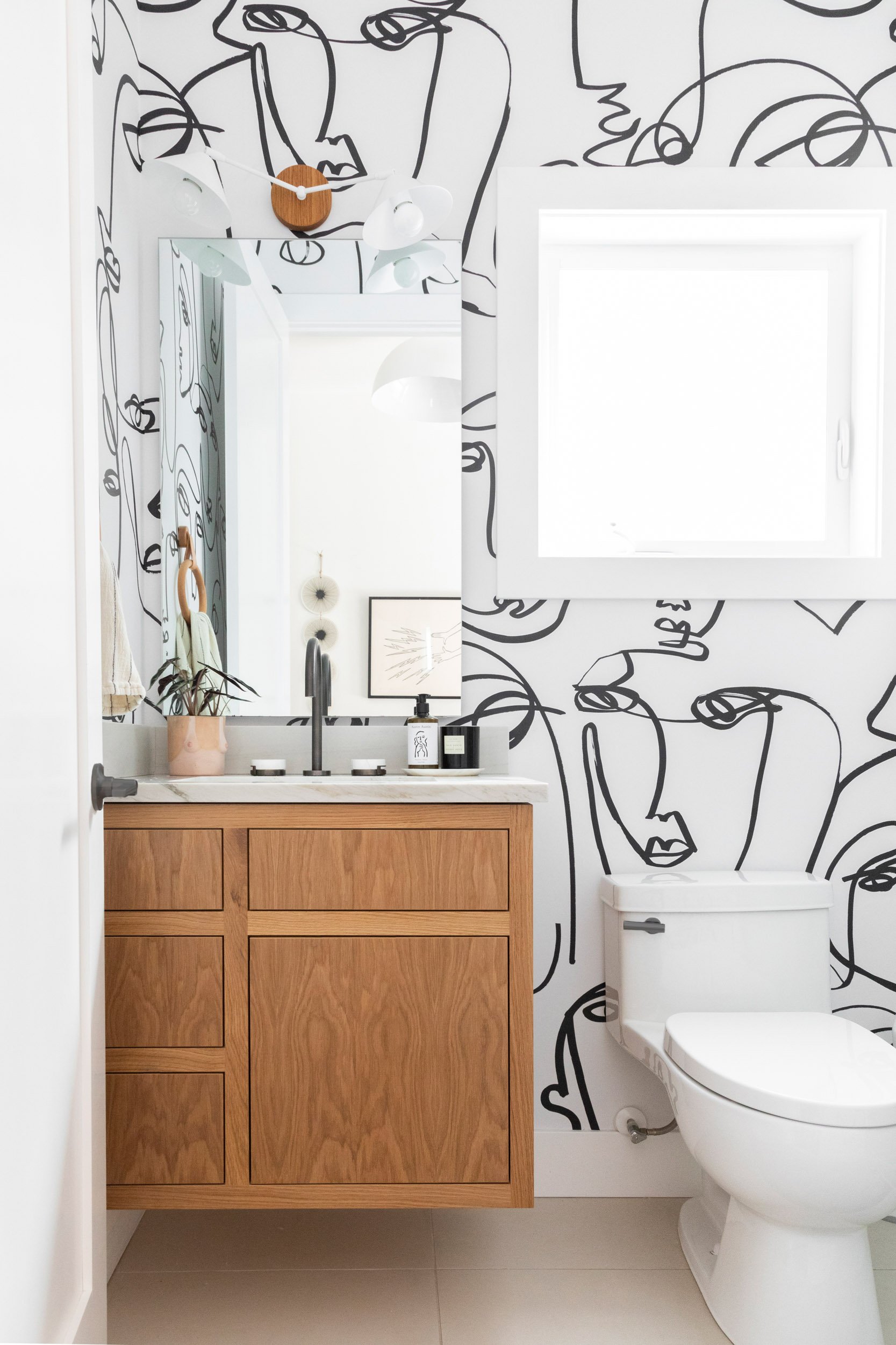 Modern bathroom with abstract line art wallpaper, wooden vanity, white toilet, large mirror, black faucet, and decor items like a plant and framed picture.