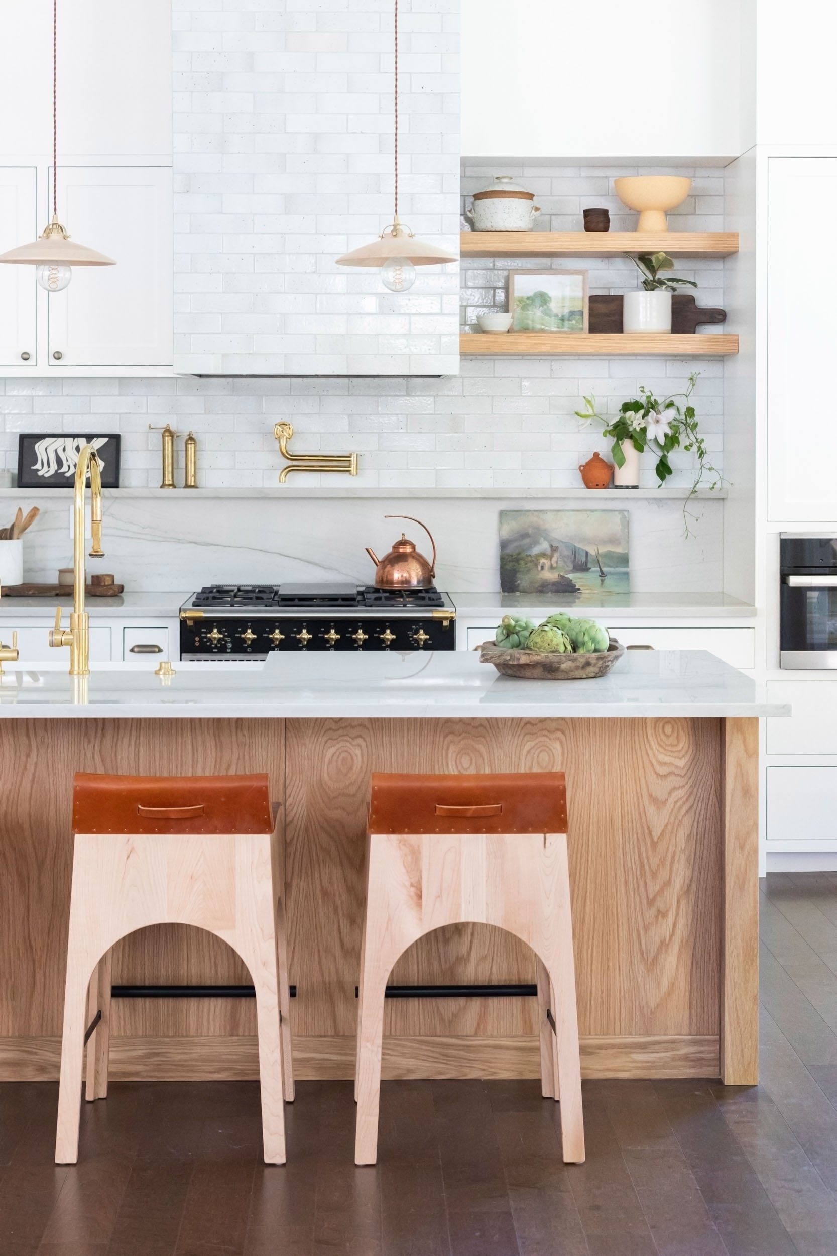 Modern kitchen with wooden island, white cabinetry, pendant lights, and decorative shelves. Includes a stove, copper kettle, plants, and artichokes in a bowl.