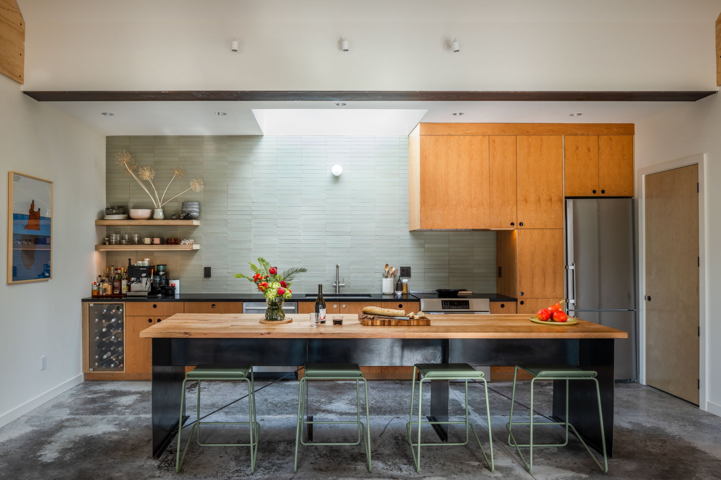 Modern kitchen with wooden cabinets, a large island with green stools, an oven, and a refrigerator. The backsplash is light gray tile, and there's a wine storage area. A vase with flowers and a bottle of wine sit on the island.