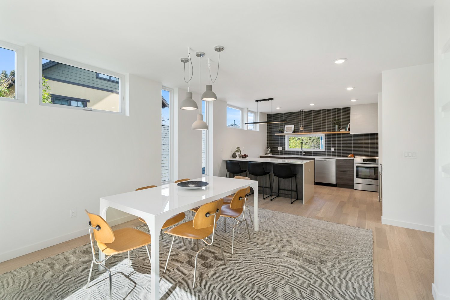 Modern open-concept dining and kitchen area with wooden chairs, a white table, and pendant lighting; features a black-tiled kitchen backsplash, island with bar stools, and stainless steel appliances.