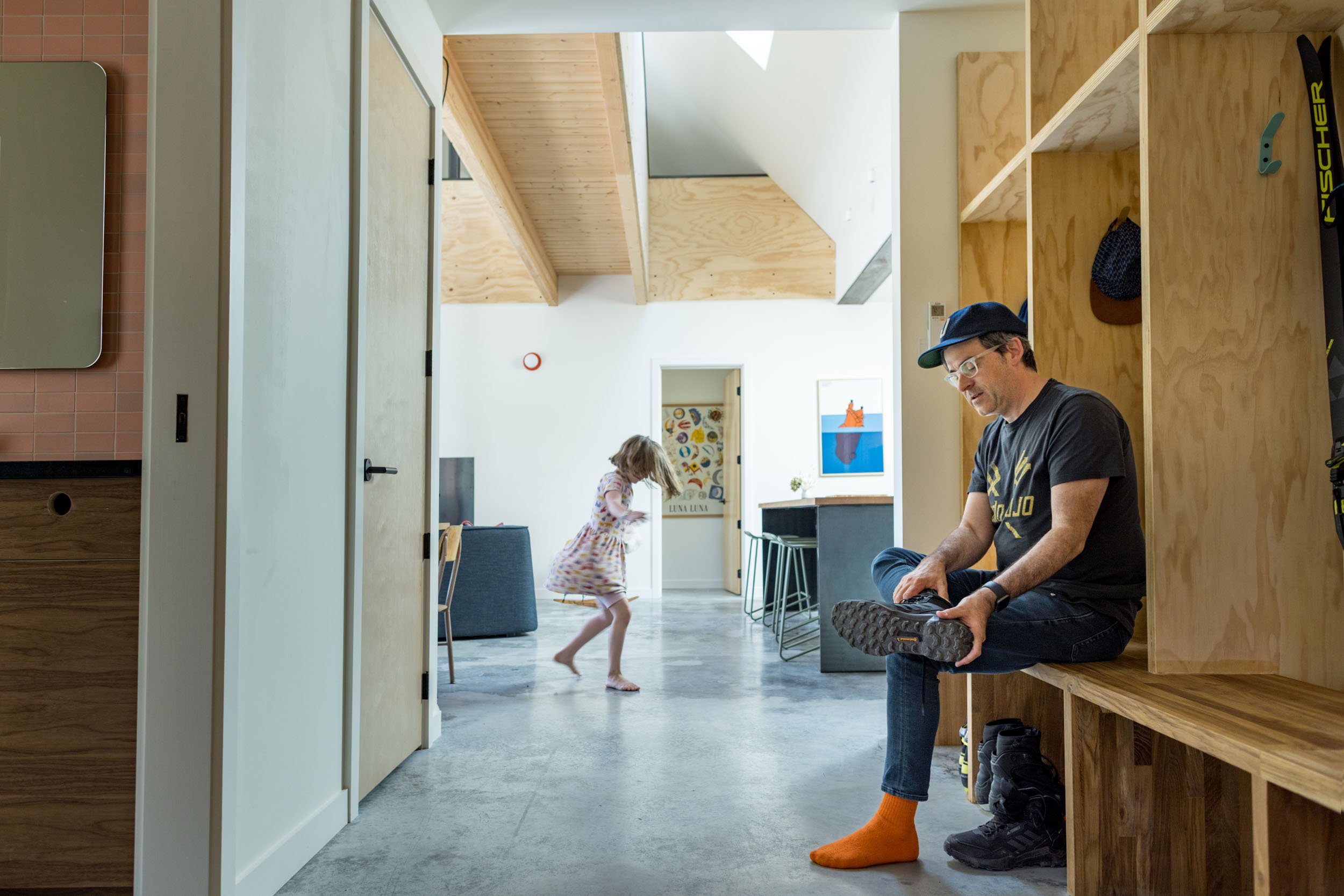 Man sitting and tying shoes in a modern home with a child running in the background.