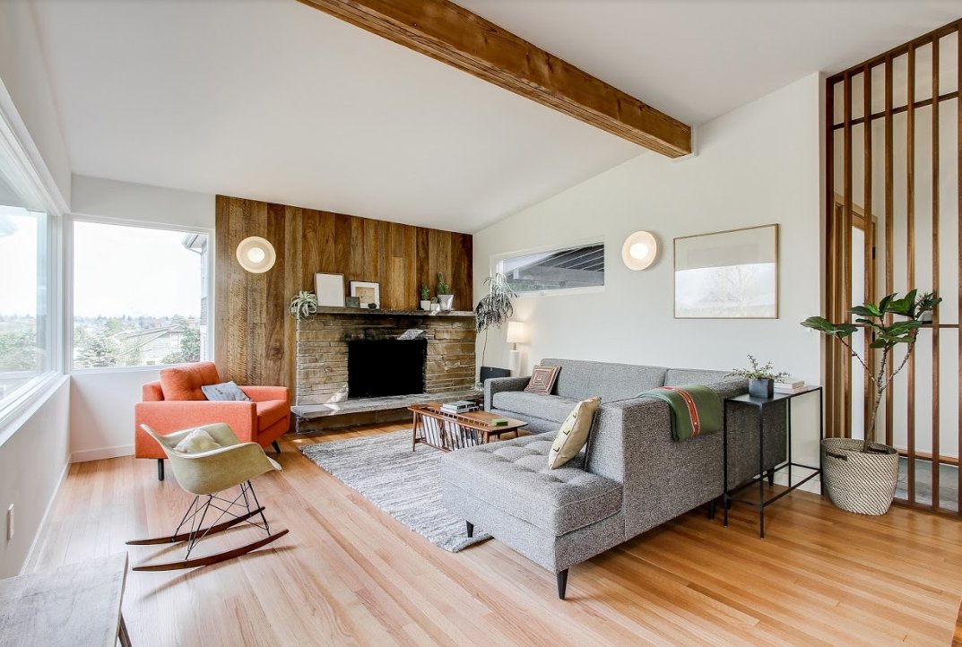 Modern living room with a gray sectional sofa, wooden accent wall, stone fireplace, orange armchair, rocking chair, plants, and decorative elements.