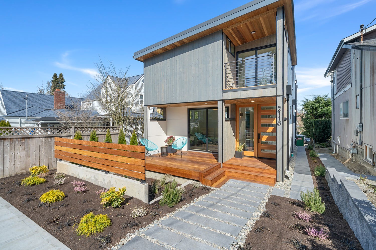 Modern two-story house with wooden exterior, small porch, and landscaped front yard with shrubs and stepping stone walkway.