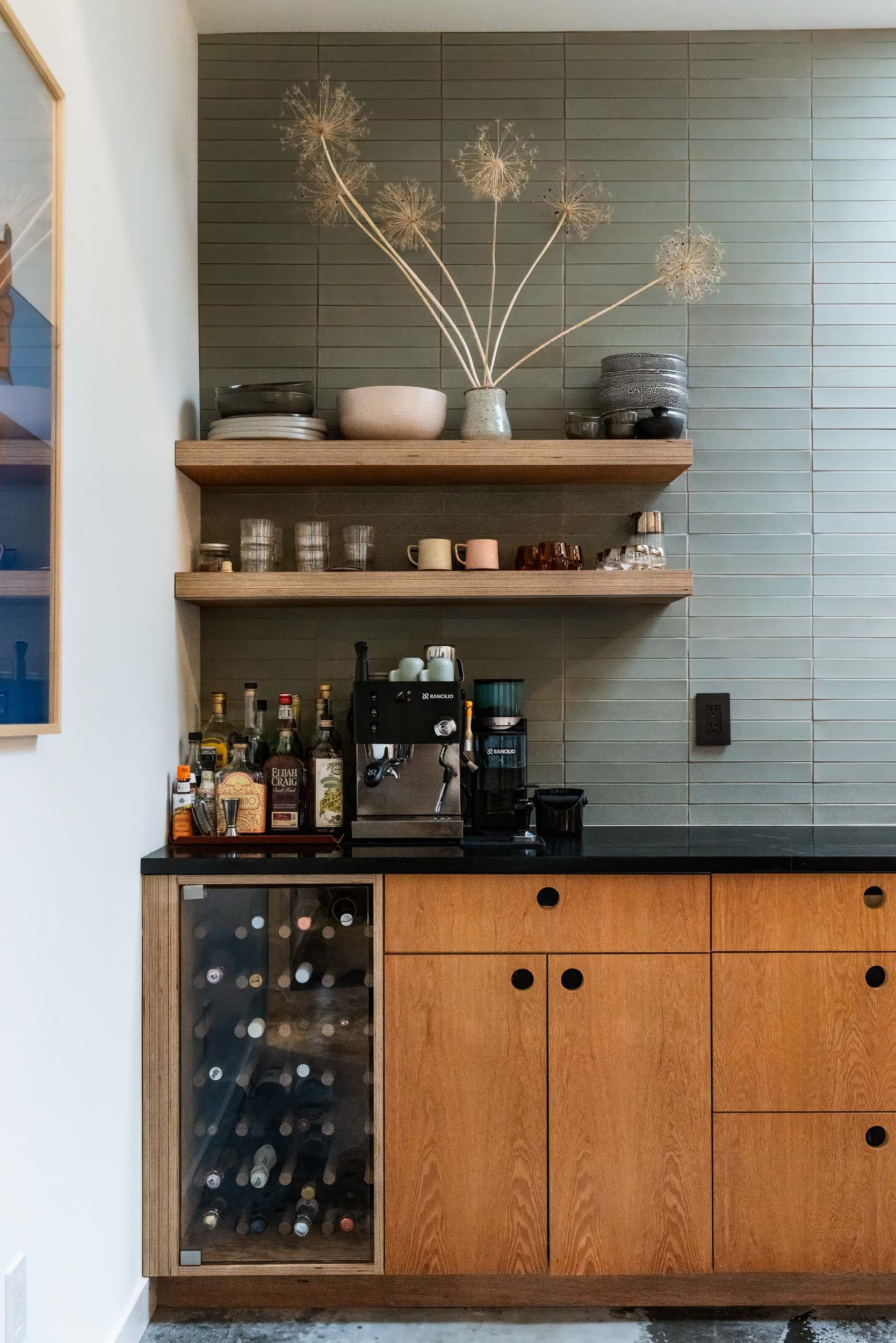 Modern home bar setup with wooden cabinets, shelves, liquor bottles, wine storage, espresso machine, coffee grinder, glassware, and decorative objects, set against a tiled wall.